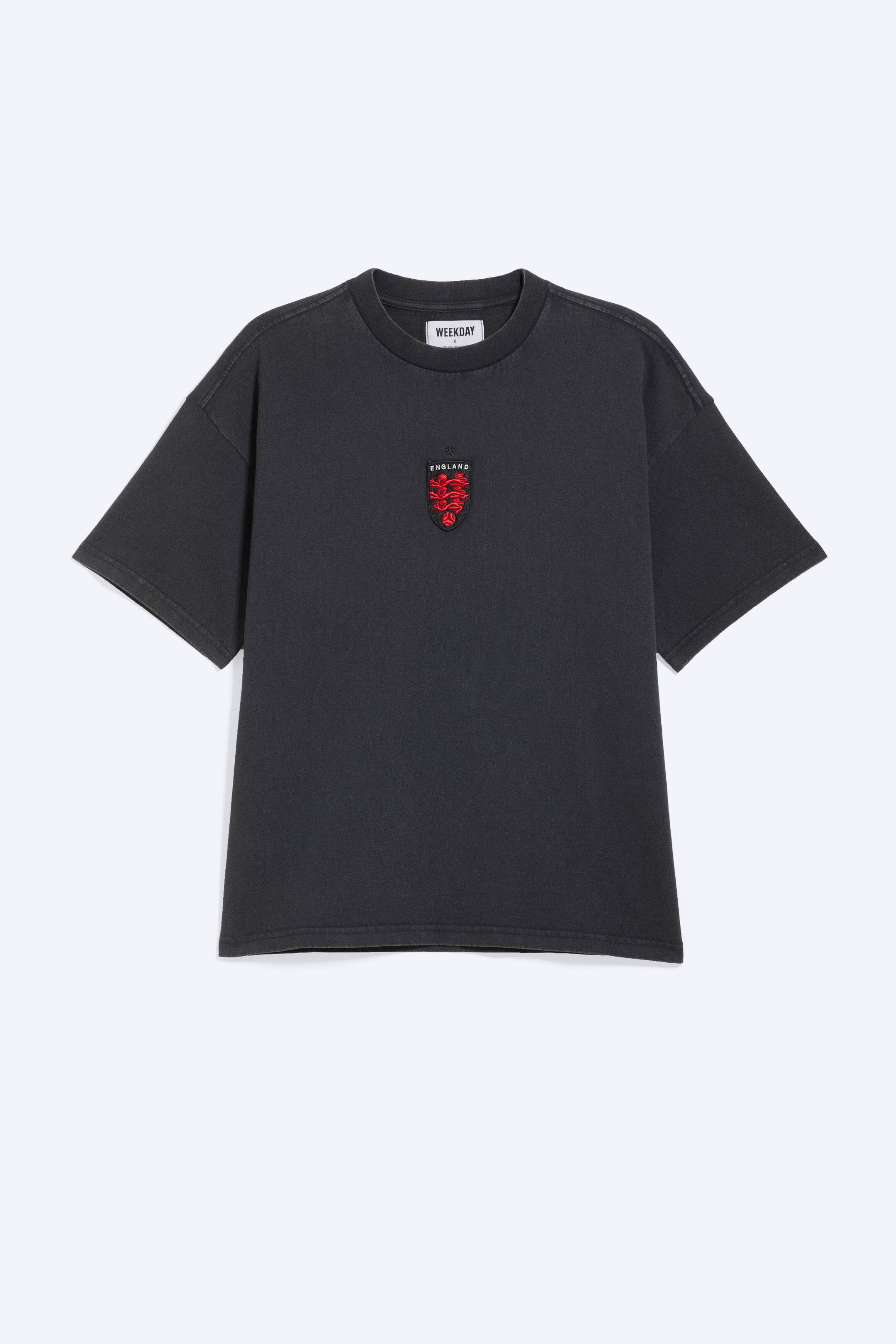 Ver imagen más grande: Se muestra una camiseta gris oscuro desde el frente, con un corte holgado, cuello redondo y mangas cortas y anchas. Un emblema rojo en forma de escudo, que presenta tres leones bordados y la palabra 'ENGLAND', está colocado de manera central en el pecho, y una etiqueta de la marca 'WEEKDAY' en color blanco es visible en el interior de la parte posterior del cuello.