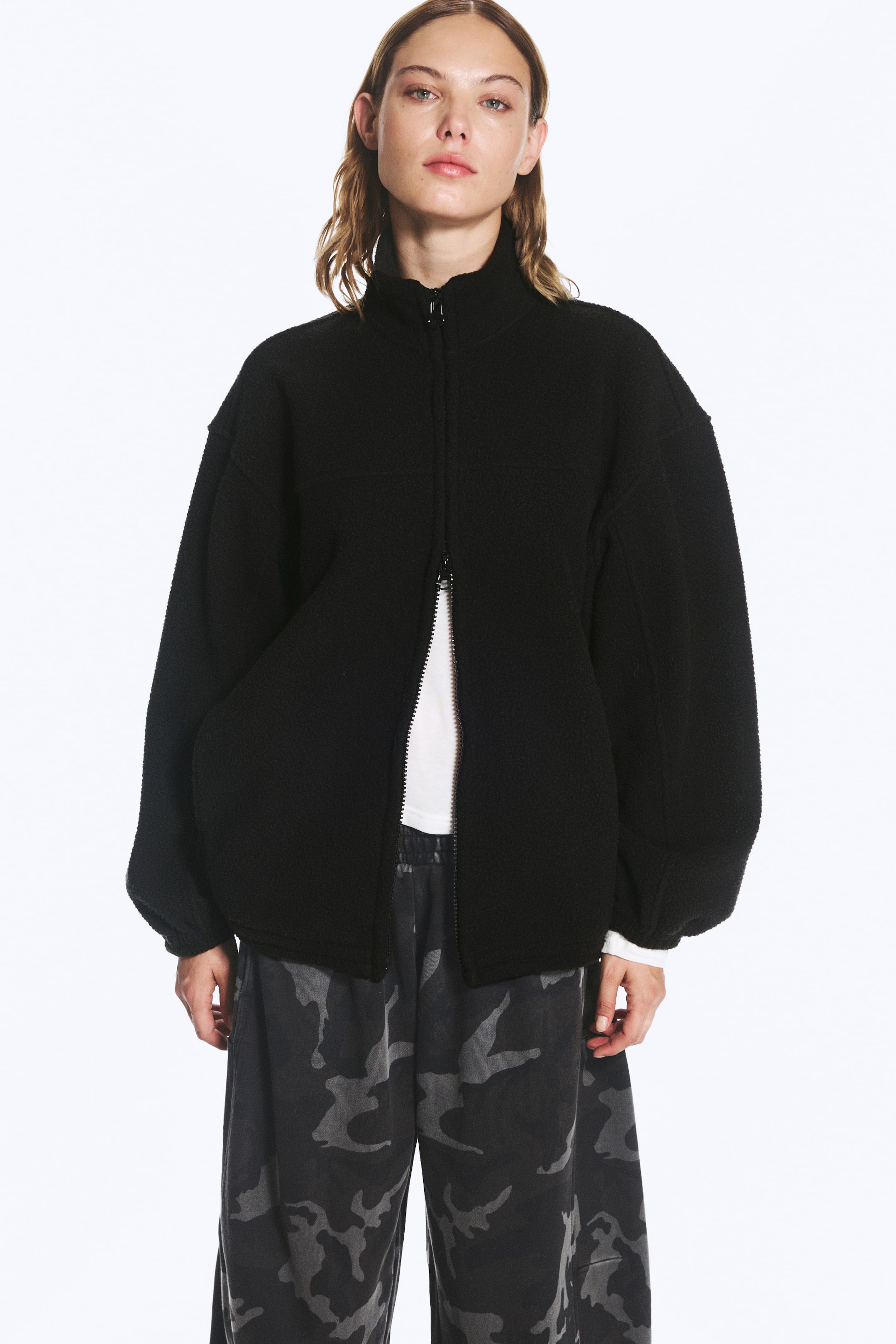 View larger image: A black fleece zip jacket featuring a high collar, dropped shoulders, and elasticated cuffs is being worn by the model. The jacket’s two-way zip is partially open at the bottom, revealing a layered white T-shirt and dark camouflage-patterned trousers.