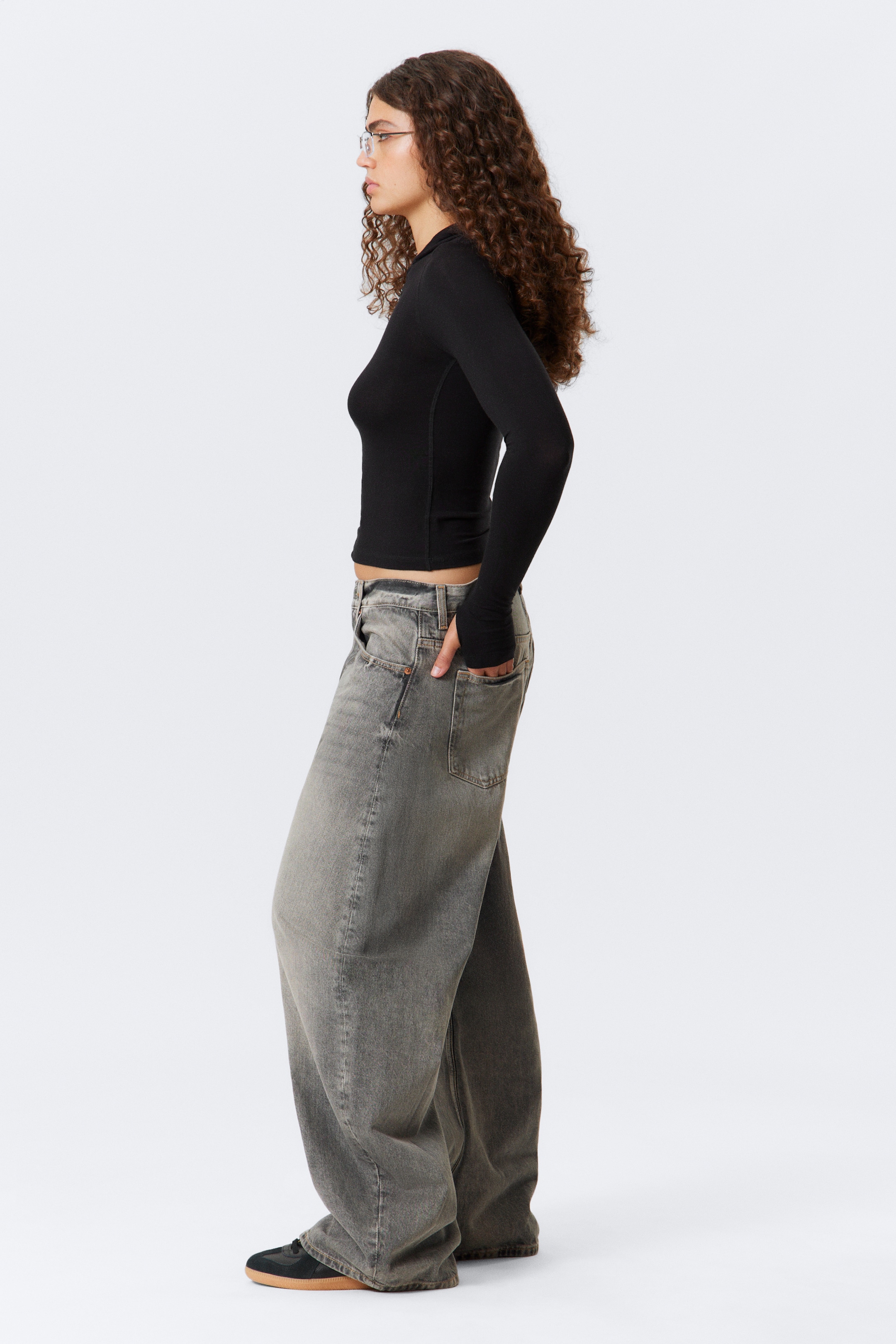 View larger image: A black, fitted, long-sleeved top with a high neckline and thumbholes is being shown from a side profile, ending above the waist. It is being worn with grey, washed wide-leg jeans featuring visible front and back pockets and slight distressing, which are pooling over black sneakers with gum soles.