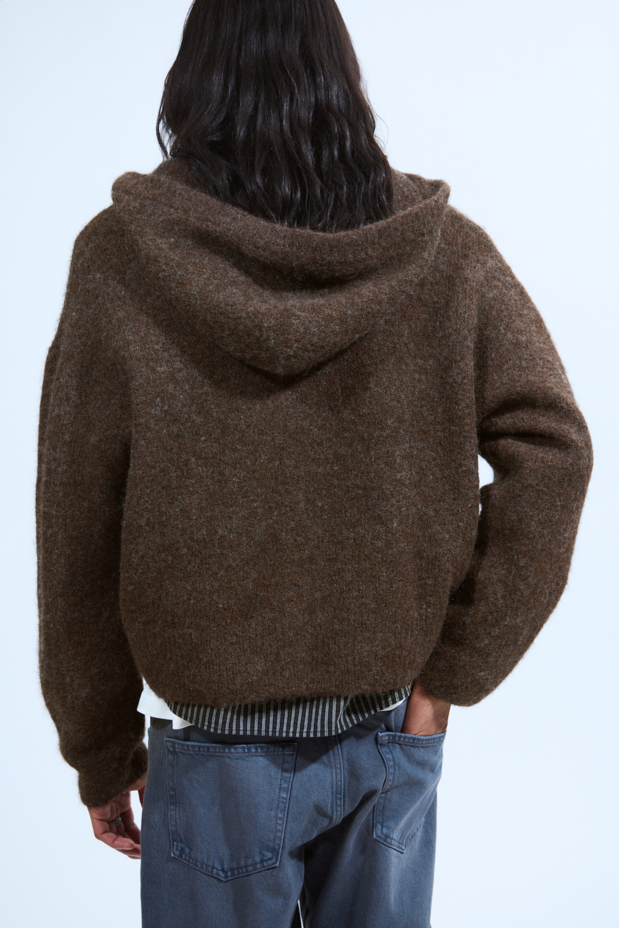 View larger image: A brown hooded sweater is being presented from the back, showcasing its fuzzy knit texture, oversized fit, and cropped hem. A striped shirt is being layered underneath the sweater.