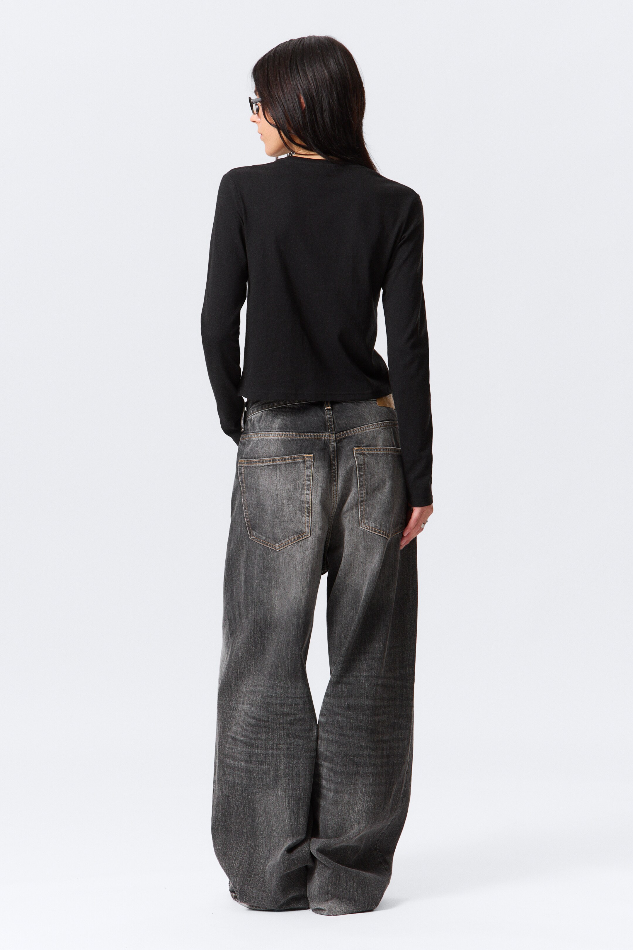 Ingrandisci l'immagine: Un top nero a maniche lunghe e jeans grigio scuro a gamba larga vengono presentati da dietro. Il top nero presenta una silhouette liscia e aderente che termina in vita, mentre i jeans grigio scuro mostrano un taglio rilassato a gamba larga con tasche posteriori visibili, passanti per cintura e una finitura lavata e consumata, inclusa una toppa in pelle marrone sulla cintura posteriore.