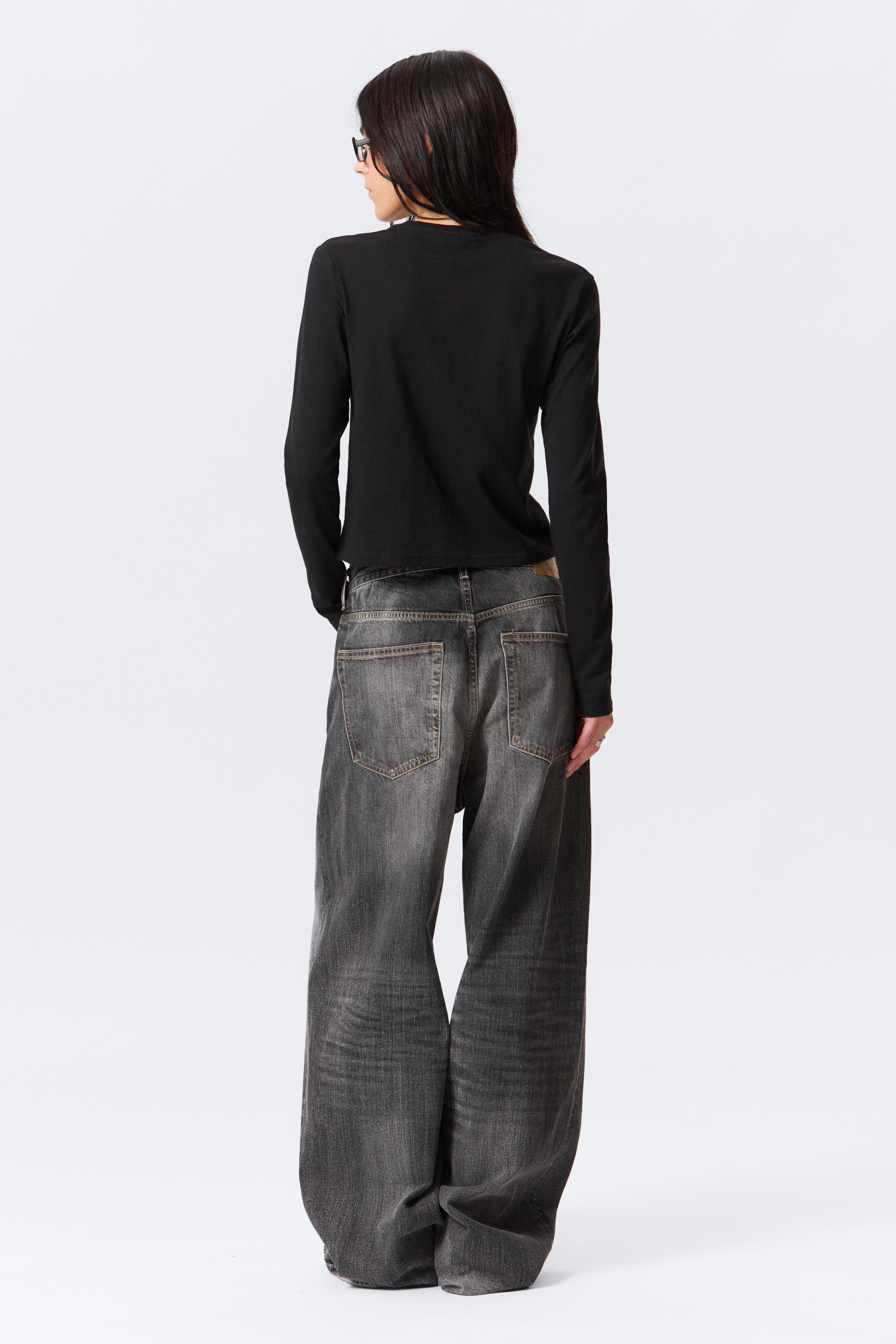 Grösseres Bild ansehen: Ein schwarzes Langarm-Top und dunkelgraue Jeans mit weitem Bein werden von hinten präsentiert. Das schwarze Top zeichnet sich durch eine glatte, figurbetonte Silhouette aus, die an der Taille endet, während die dunkelgrauen Jeans einen entspannten, weiten Schnitt mit sichtbaren Gesäßtaschen, Gürtelschlaufen und einem abgenutzten Wasch-Finish aufweisen, einschließlich eines braunen Lederaufnähers am hinteren Bund.