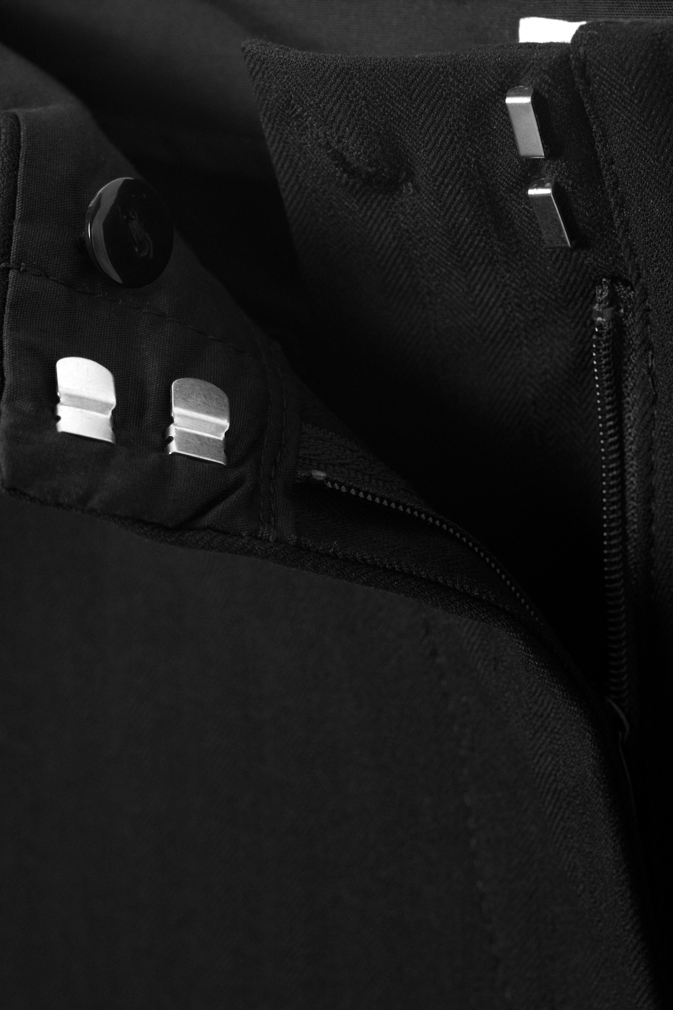 Grösseres Bild ansehen: Schwarze Hosen verfügen über ein Verschlusssystem an der Vorderseite, bestehend aus einem schwarzen Knopf, zwei silberfarbenen Haken- und Riegelverschlüssen und einem teilweise offenen Reissverschluss.