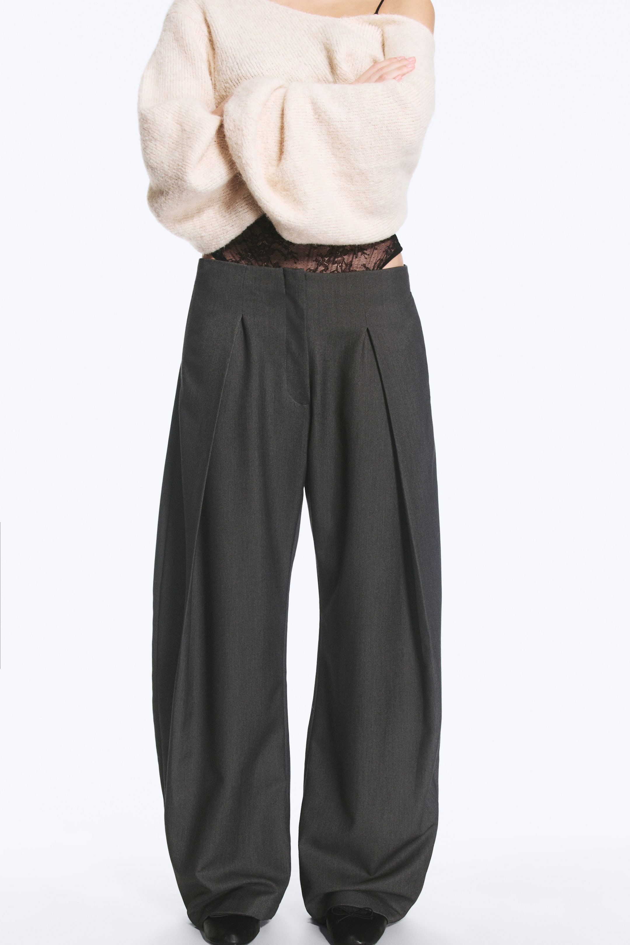 Ver imagen más grande: La modelo lleva unos pantalones globo de tiro bajo en gris oscuro, con pinzas profundas en la parte delantera y una silueta voluminosa de pierna ancha. Los pantalones se combinan con un jersey de punto en color crema y una capa de encaje negro visible en la cintura de tiro bajo.