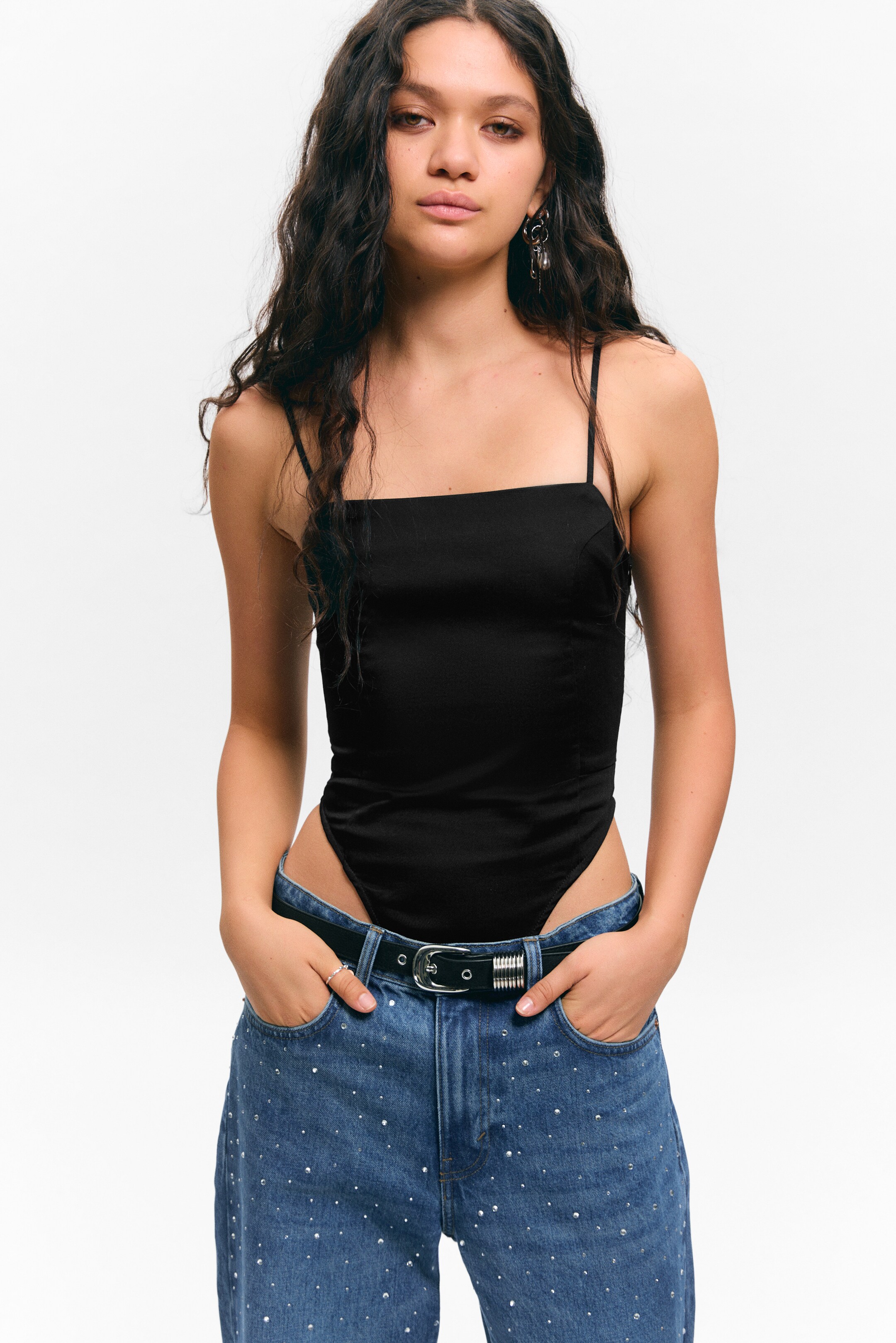 Agrandir l'image: Un bodysuit noir fait face vers l'avant, avec un décolleté carré et de fines bretelles spaghetti. Fabriqué dans un tissu noir lisse, le vêtement ajusté présente une découpe haute de la jambe où il est rentré dans un jean bleu.