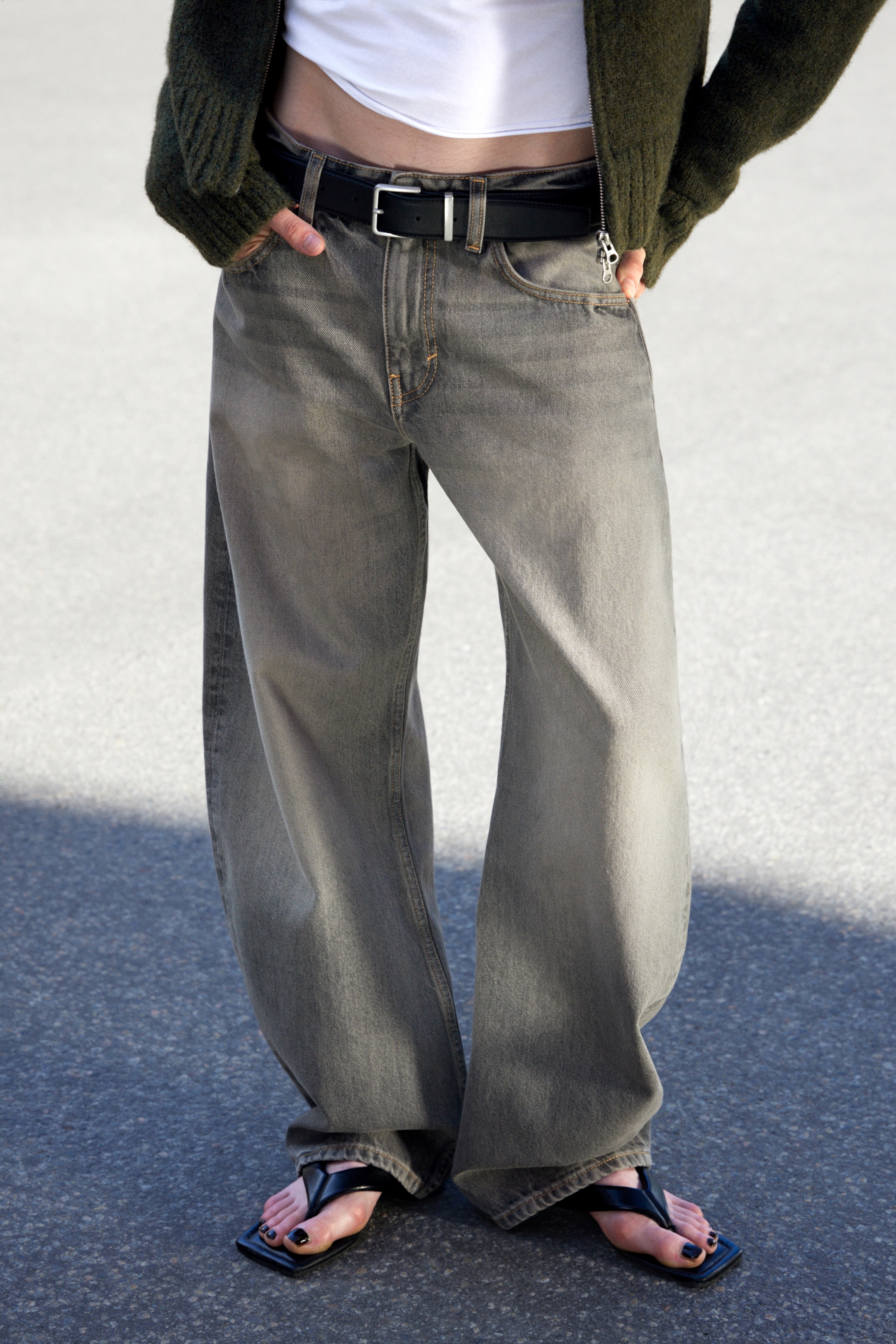 Grösseres Bild ansehen: Dunkelgraue, weit geschnittene Jeans werden in einer frontalen Ansicht präsentiert, mit einer entspannten Passform und sichtbaren kontrastierenden braunen Nähten. Sie werden hoch tailliert mit einem schwarzen Gürtel getragen und sammeln sich an den Knöcheln über schwarzen Sandalen mit quadratischer Spitze.