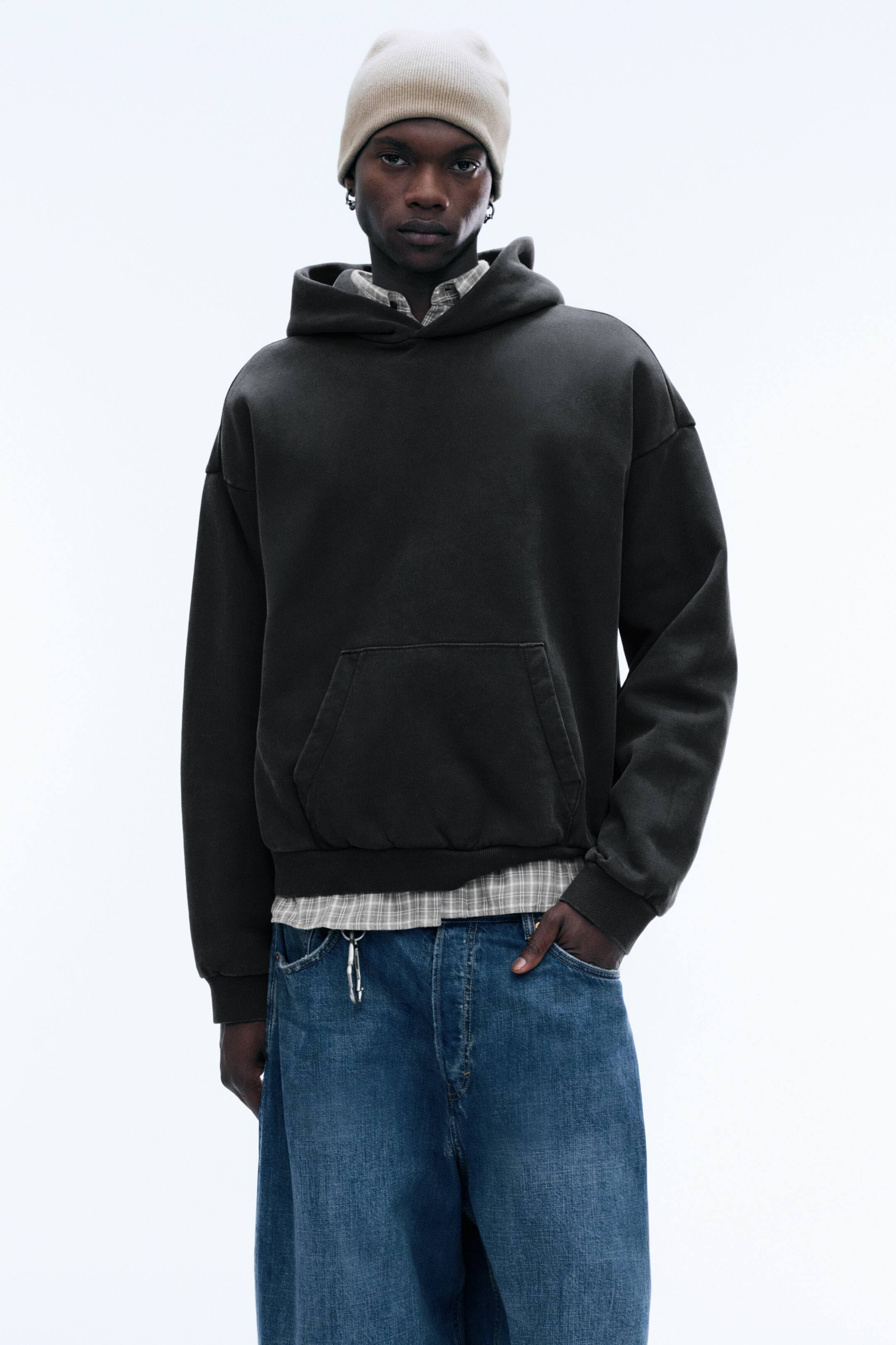 Größeres Bild anzeigen: Ein dunkelgrauer Hoodie in lockerer Passform wird vom Model getragen und verfügt über eine strukturierte Kapuze ohne Kordelzug, überschnittene Schultern und eine Kängurutasche vorne. Der Hoodie wird über einem hellen karierten Hemd getragen und mit einer blauen Jeans sowie einer beigen Beanie kombiniert.