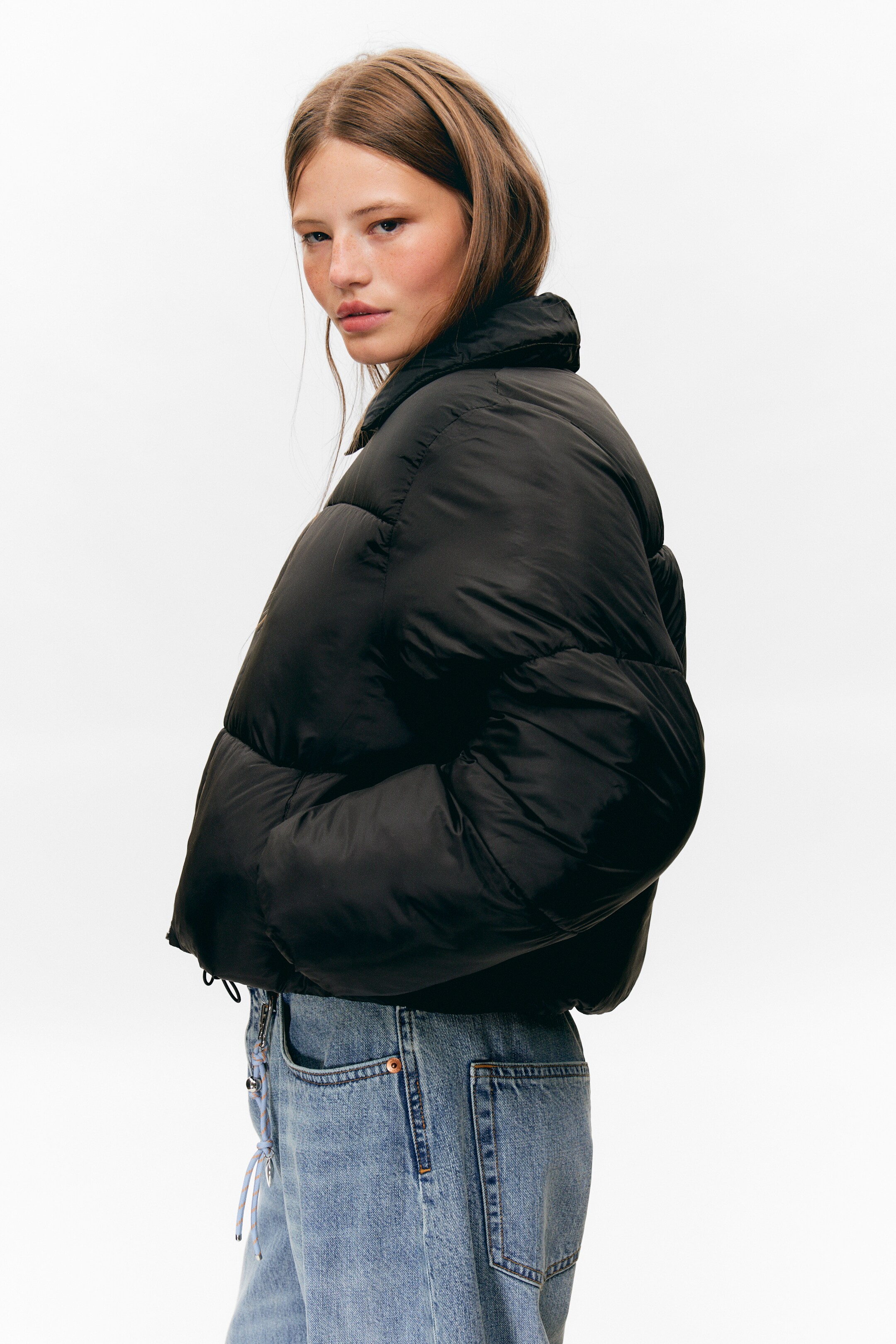 Visa större bild: En svart puffjacka presenteras från en sidobakre vinkel, vilket visar upp dess voluminösa, korta design. Den har en hög ståkrage och en slät, något glansig yta med vadderad konstruktion, medan dess fåll är åtdragen med en synlig dragsko.