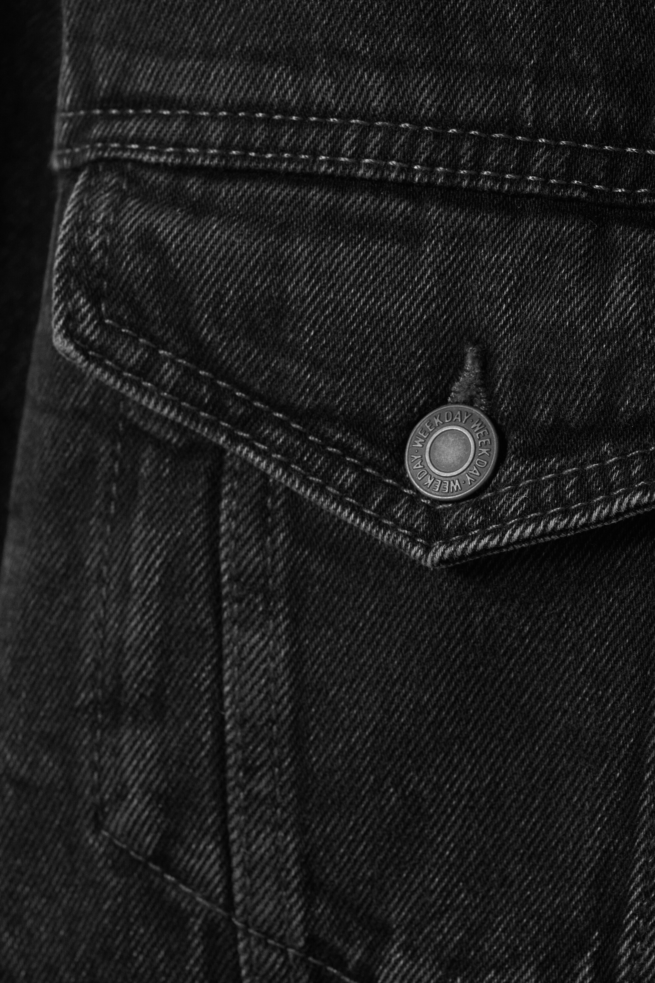 View larger image: A black denim jacket is showing a close-up of its front chest pocket, which is featuring a flap. The flap is secured by a round, dark metal button embossed with 'WEEKDAY' text, and light-colored stitching is detailing the pocket and surrounding seams.