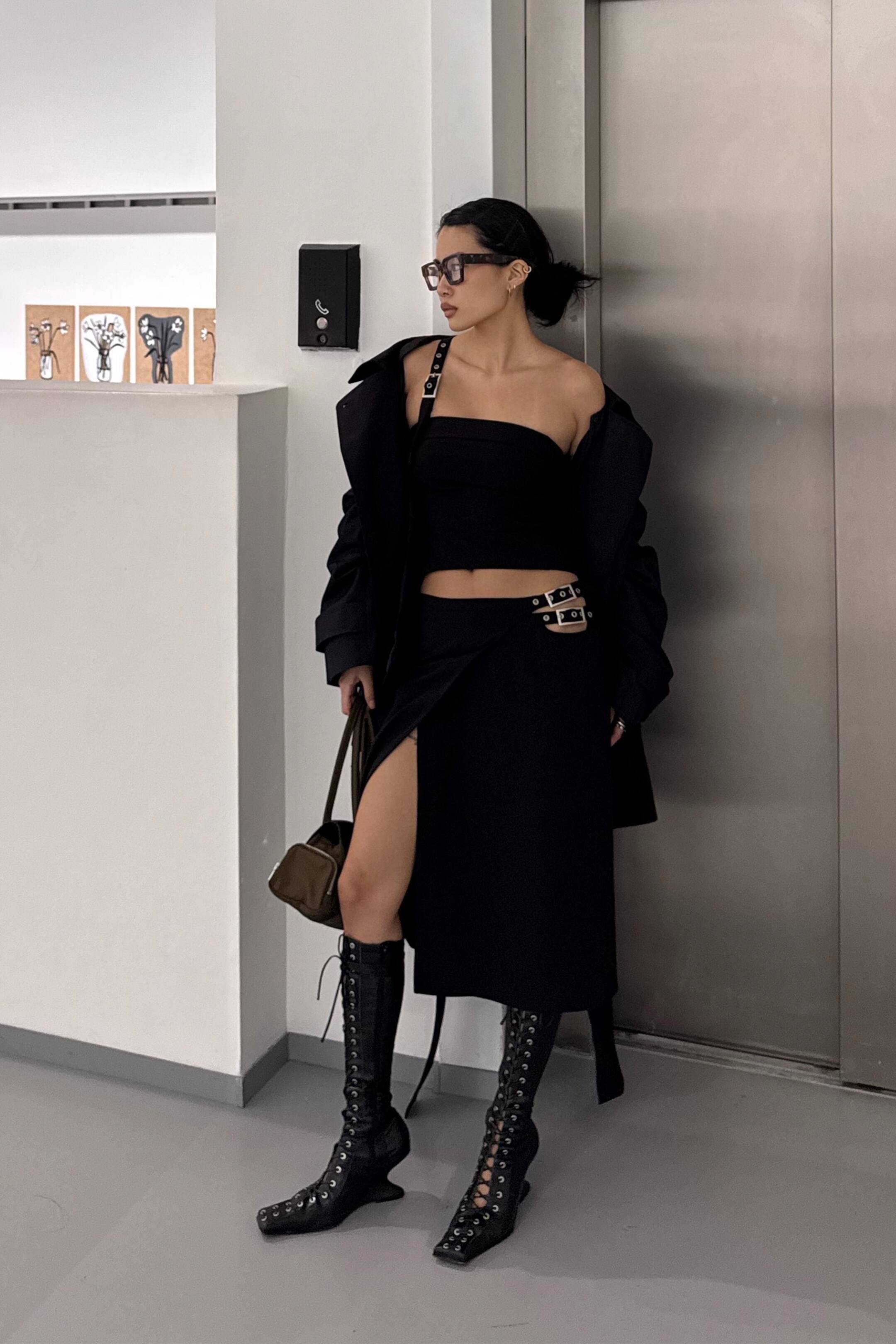 Nagyobb kép megtekintése: Egy fekete midi szoknya, amely elöl átkötős stílusban van kialakítva, magas bal oldali sliccel. Két téglalap alakú, ezüst színű csat rögzíti az átfedő anyagot a csípőnél, miközben a szoknya éppen a derék alatt helyezkedik el.