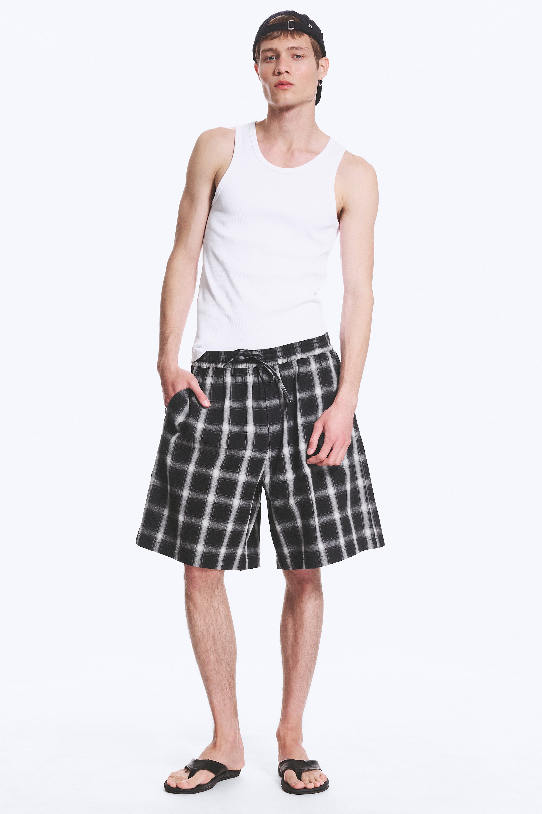 Grotere afbeelding bekijken: Zwart-wit geruite pull-on shorts worden gedragen door het model, met een losse pasvorm, een lengte tot halverwege het dijbeen en een elastische tailleband met een zichtbare trekkoordknoop. Het model heeft één hand in een zak van de shorts gestoken.