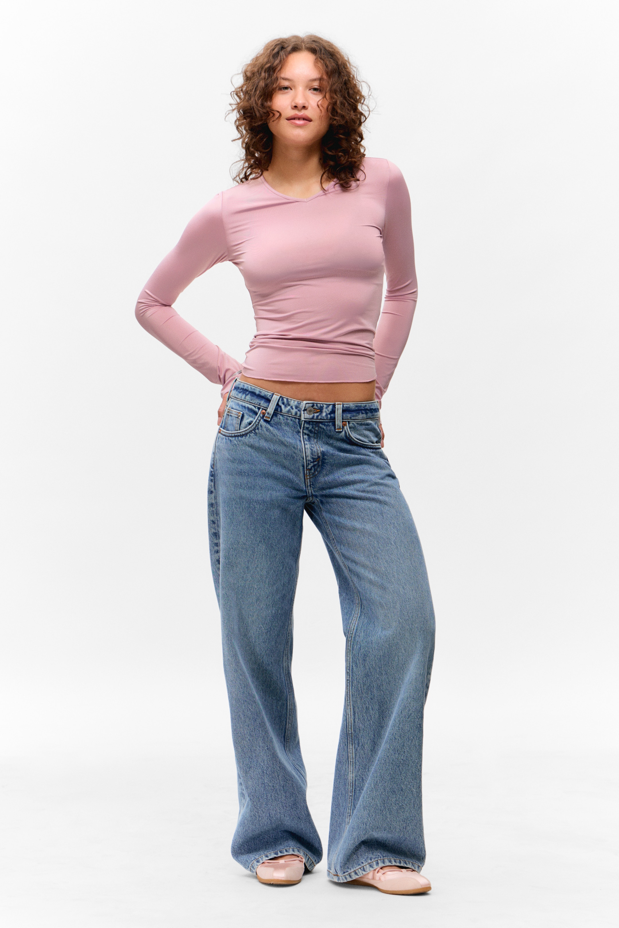 Größeres Bild ansehen: Ein hellrosa, langärmliges Oberteil mit V-Ausschnitt wird getragen, es sitzt eng an und endet knapp über einer hellblauen, weit geschnittenen Jeans. Die Jeans sitzen in mittlerer Höhe, und rosa Lackleder-Ballerinas vervollständigen das nach vorne gerichtete Ensemble.
