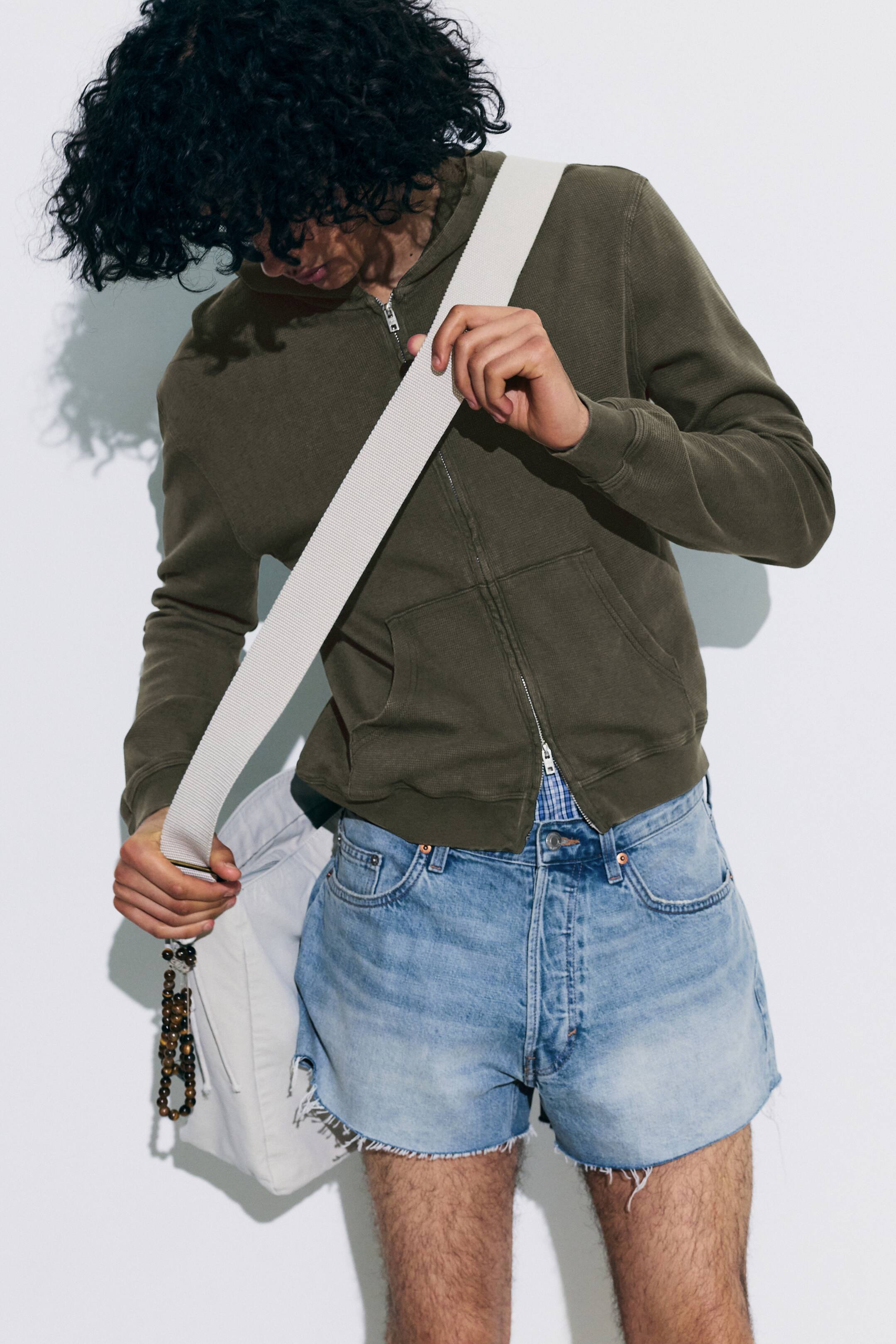 Agrandir l'image: Sweat à capuche zippé intégral vert foncé, confectionné dans un tissu tricoté texturé, avec une fermeture éclair argentée courant le long de l'avant et des poches kangourou. Les poignets et l'ourlet côtelés sont visibles, et une chemise à carreaux bleue et blanche dépasse en dessous.