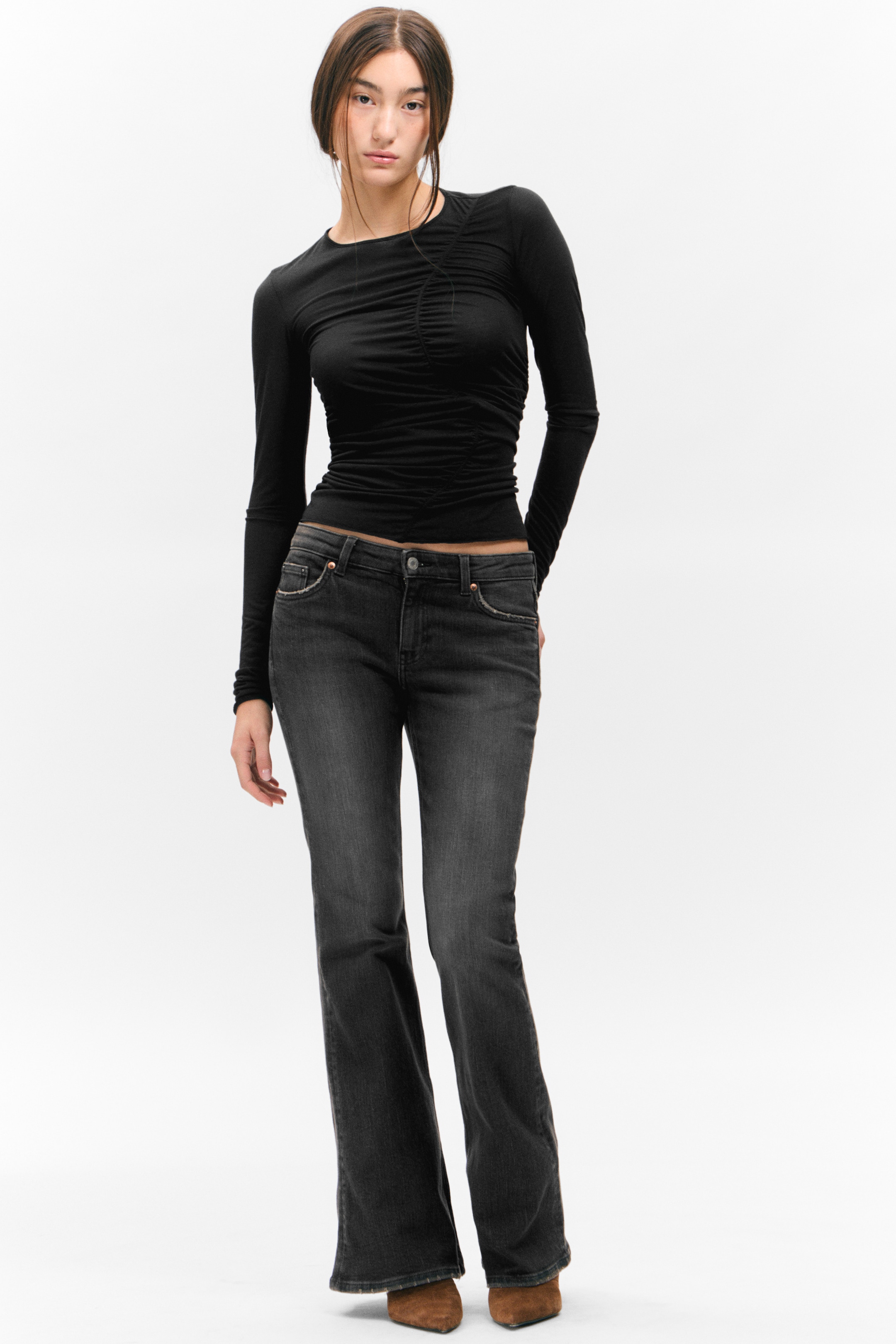Visualizza immagine più grande: Un top nero a maniche lunghe, con arricciature raccolte sul davanti e sul busto, viene abbinato a jeans bootcut grigio scuro, con un lavaggio sbiadito e tasche anteriori. Questo completo è presentato frontale, con scarpe a punta marroni visibili sotto i jeans.
