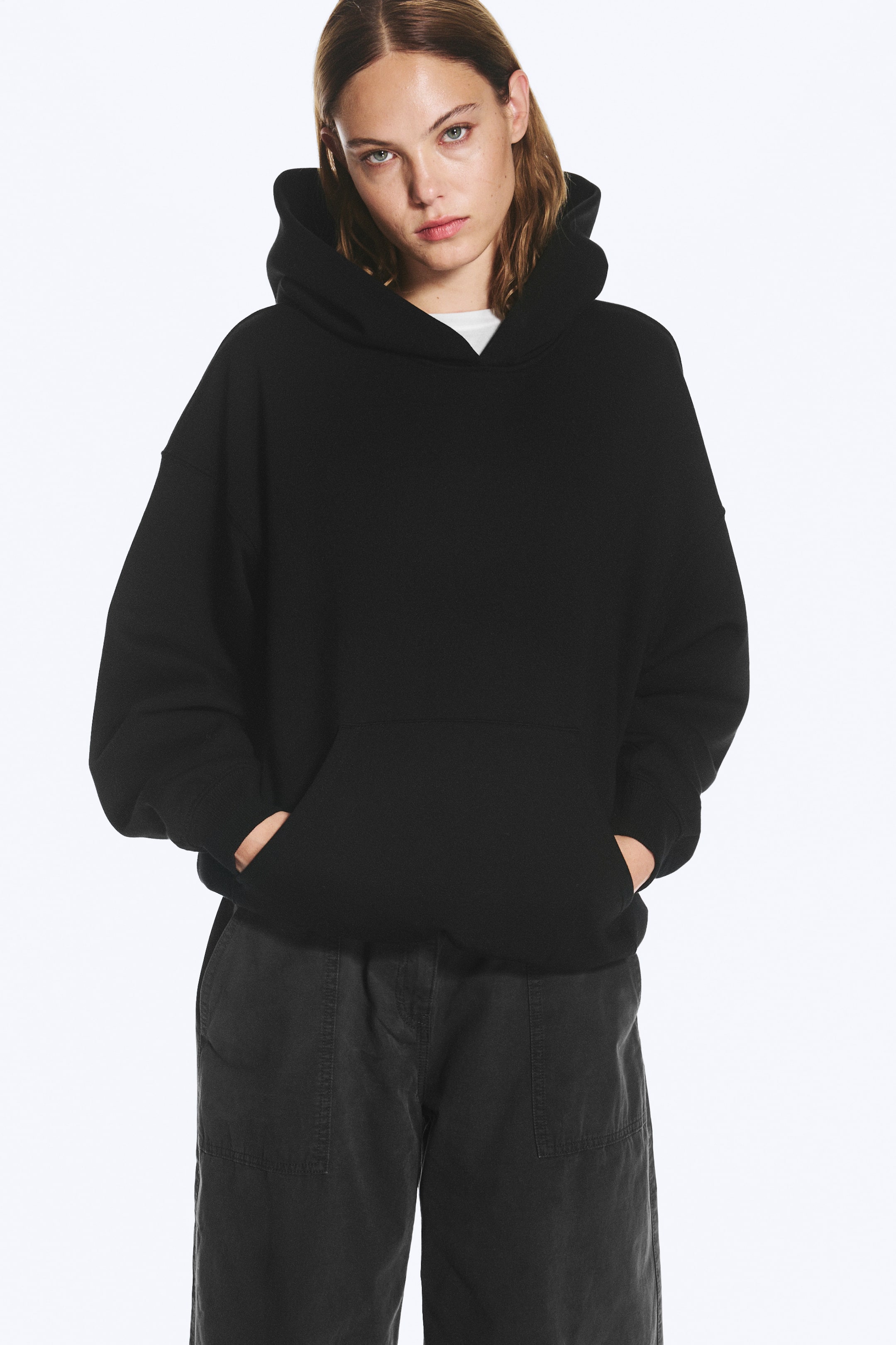 Προβολή μεγαλύτερης εικόνας: Ένα μαύρο oversized φούτερ με κουκούλα φοριέται από το μοντέλο, το οποίο στέκεται με τα δύο χέρια χωμένα στην μπροστινή τσέπη καγκουρό. Το φούτερ διαθέτει χαλαρή σιλουέτα με πεσμένους ώμους, ριμπ μανσέτες και μεγάλη κουκούλα, φορεμένο πάνω από λευκό t-shirt και σκούρο γκρι παντελόνι.