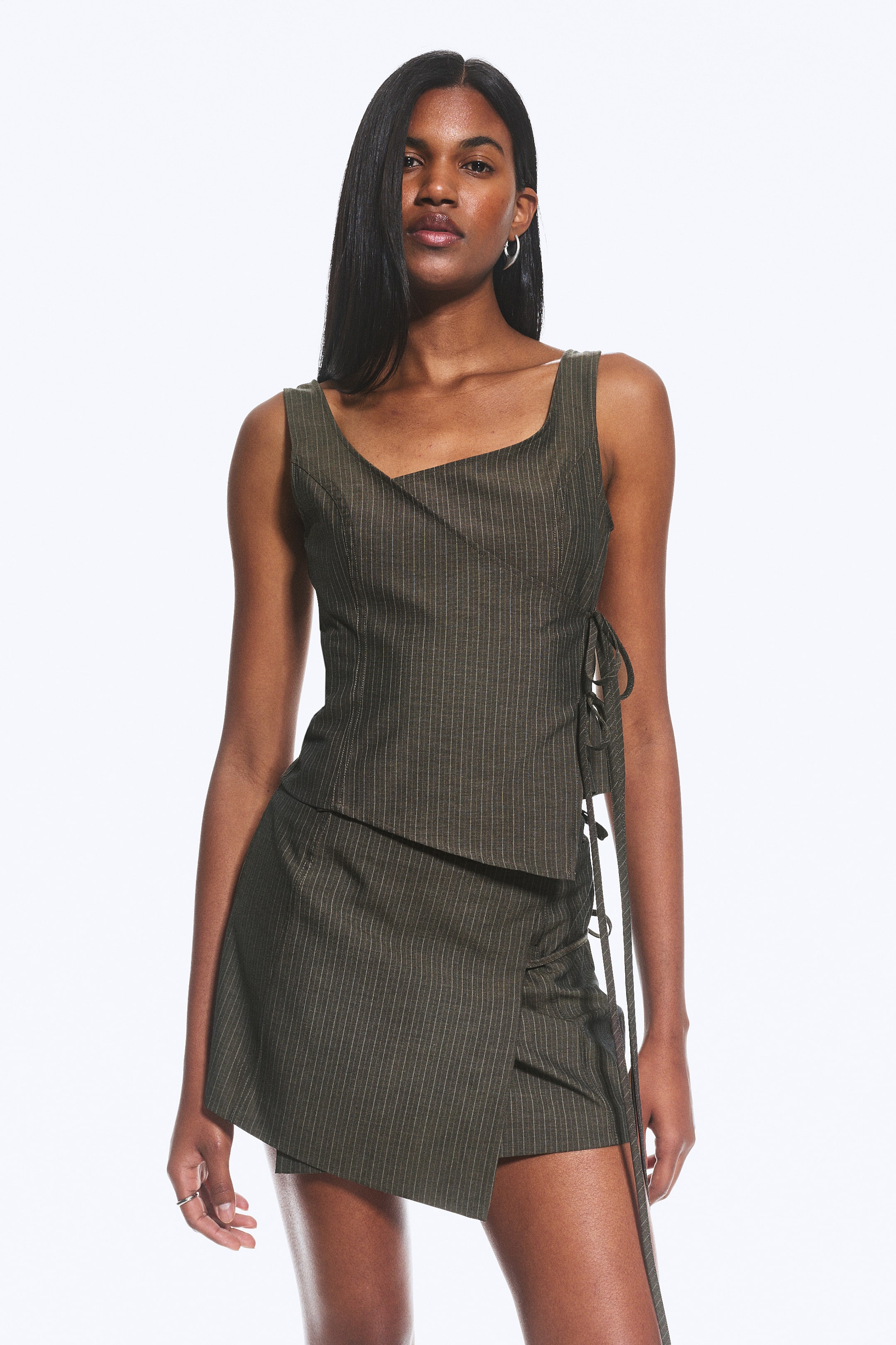 View larger image: A dark green pinstriped sleeveless top featuring an asymmetrical neckline and side lacing, viewed from a front-facing angle. It is paired with a matching pinstriped asymmetrical mini skirt, which displays a wrap-style front and side gathering.