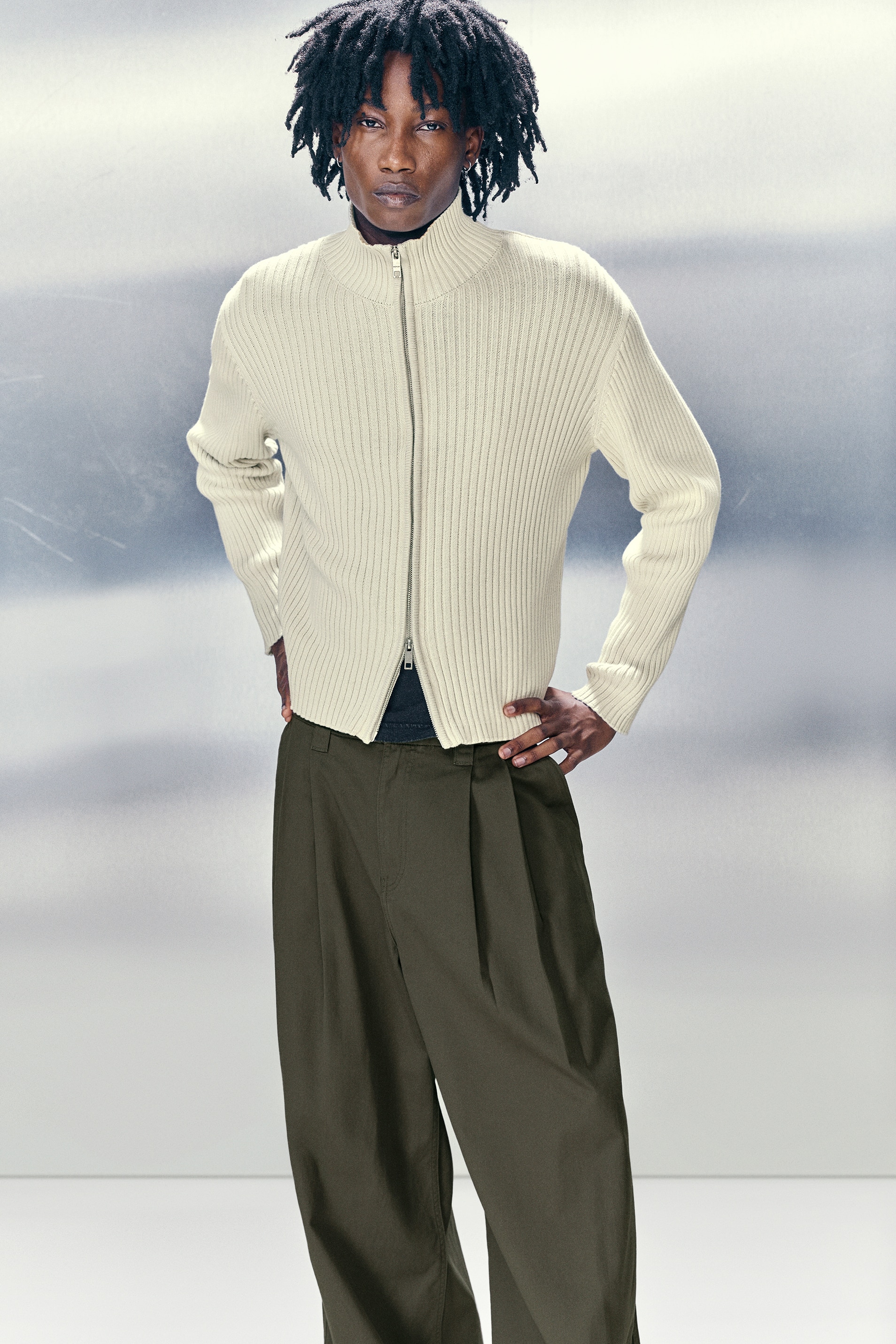 Ein hellfarbener, locker sitzender Cardigan wird von vorne präsentiert. Er zeichnet sich durch eine gerippte Strickstruktur, einen hohen Kragen und einen Zwei-Wege-Reißverschluss an der Vorderseite aus.
