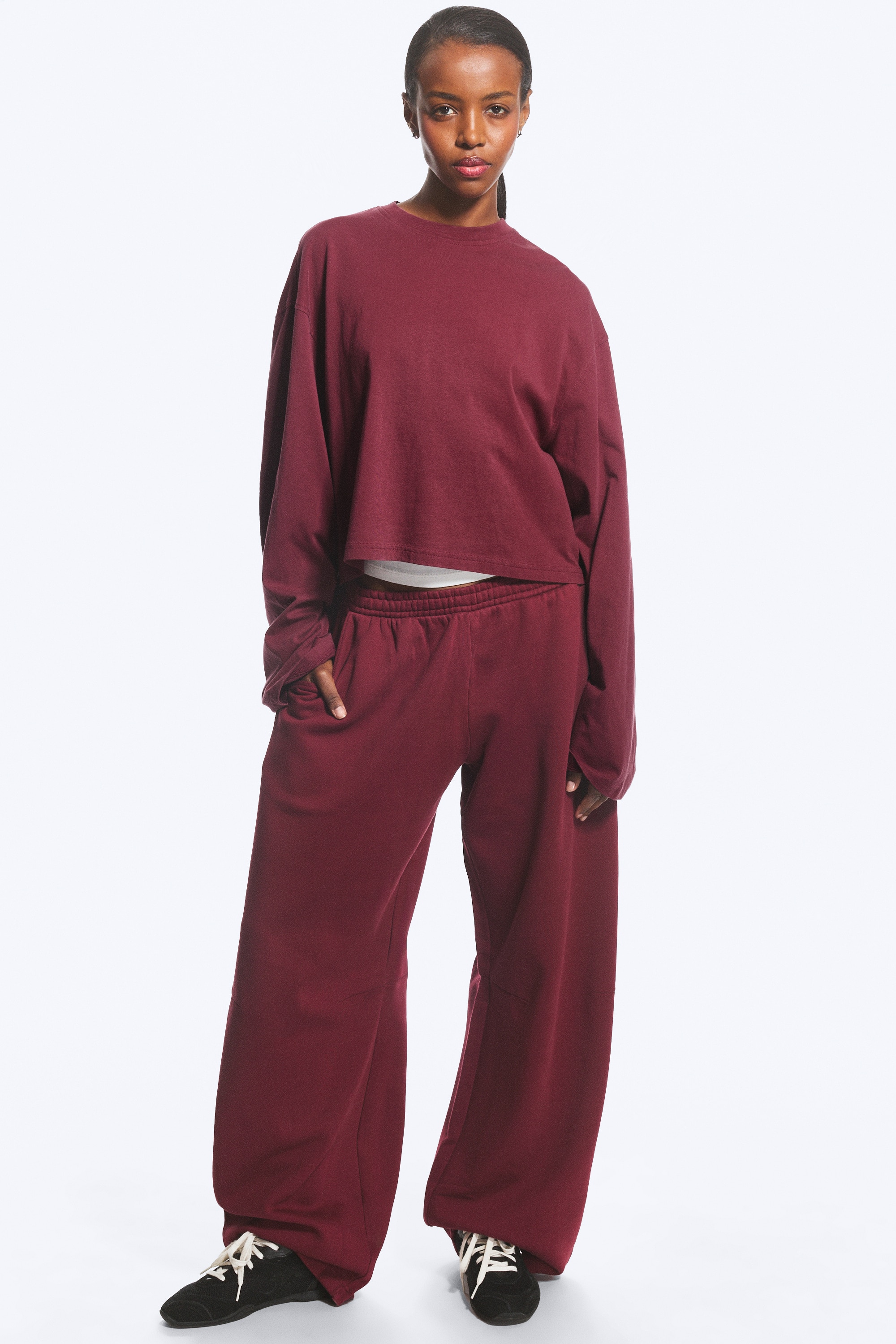 Een paar bordeauxrode sweatpants met losse pasvorm is van voren weergegeven, met een elastische tailleband en schuine steekzakken aan de zijkant. De broek heeft een wijde, barrel-vormige pijp die naar buiten toe uitloopt, met horizontale vormnaden ter hoogte van de knieÃ«n.