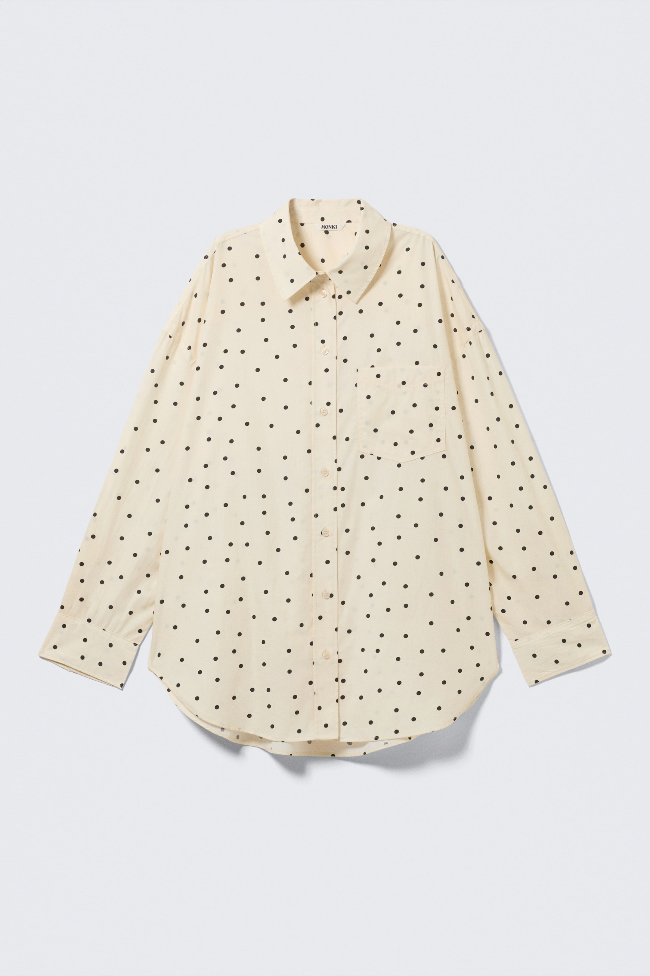 Visualizza immagine più grande: Una camicia oversize color crema a maniche lunghe con pois neri è presentata frontalmente. Presenta un colletto classico, una chiusura frontale con bottoni, una tasca singola sul petto, polsini con bottoni e un orlo curvo.