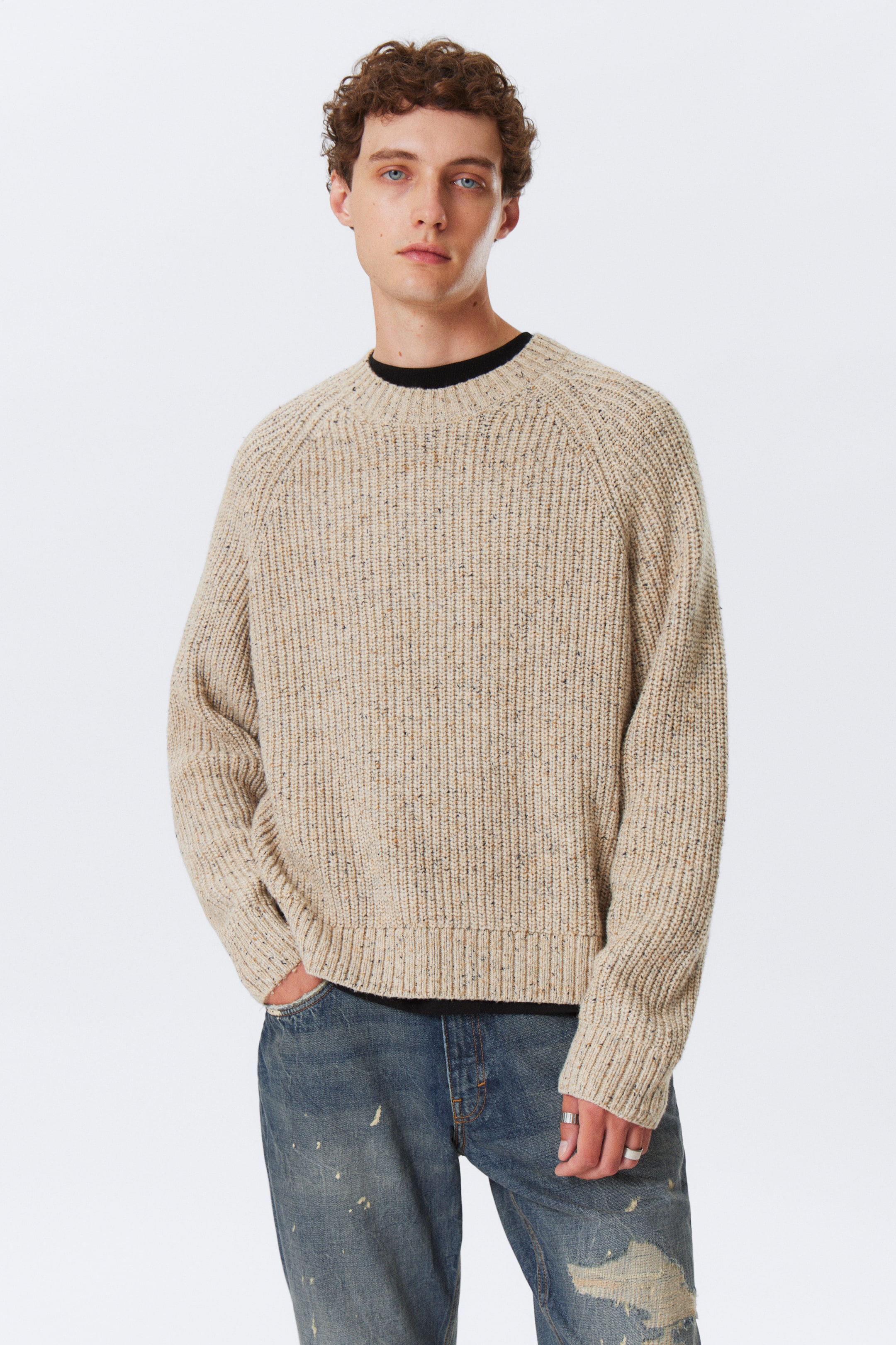 Ver imagen más grande: Se presenta un suéter de punto Donegal beige, con prominentes costillas verticales. Está diseñado con un cuello redondo, mangas raglán y terminado con puños y dobladillo acanalados, con una camiseta negra visible debajo del escote del suéter.