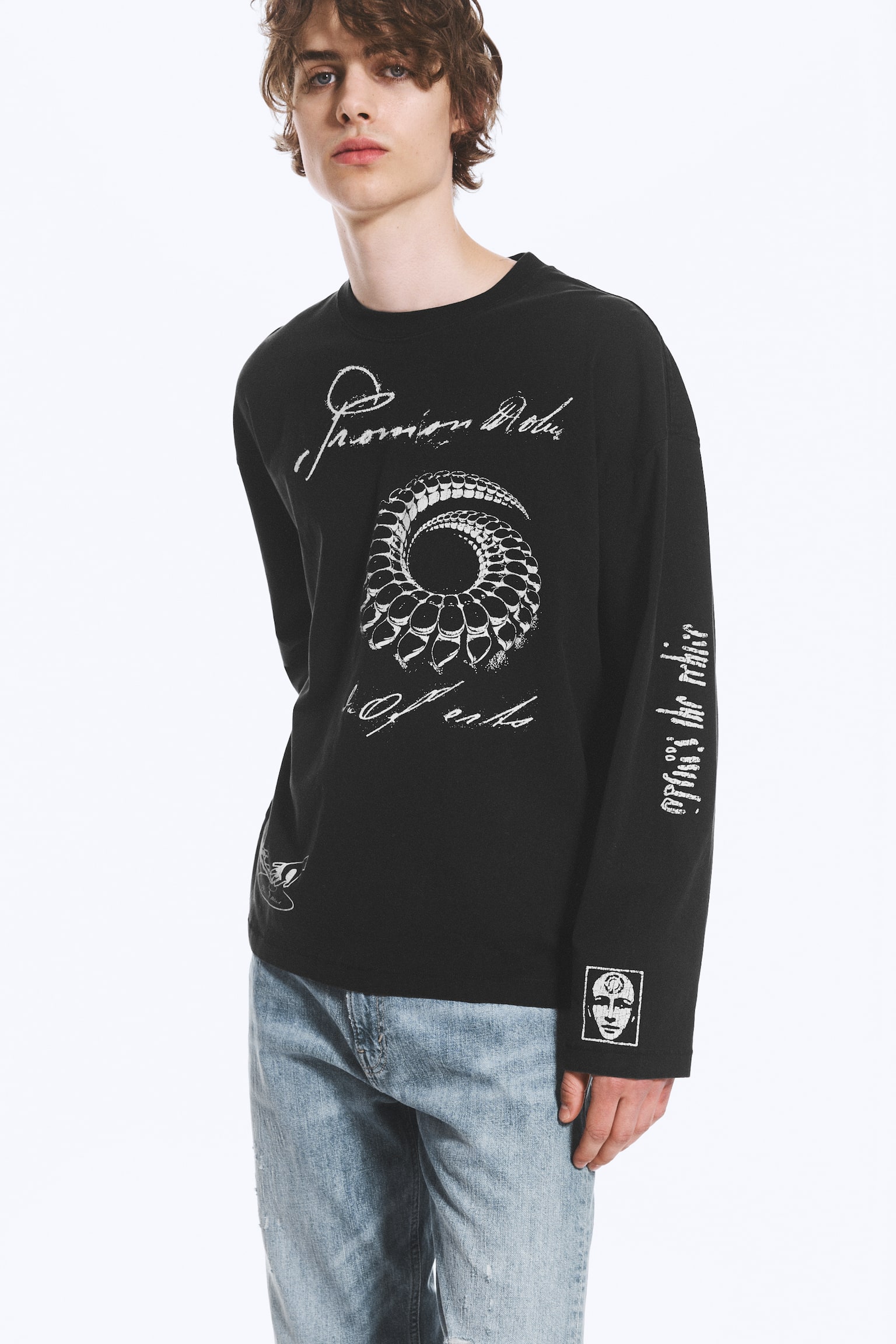 En svart långärmad t-shirt med en stor vit spiraldesign och vit skripttext på framsidan. Vit skripttext löper vertikalt på vänster ärm, och en liten fyrkantig grafik som avbildar ett ansikte finns på höger ärmslut. - 1