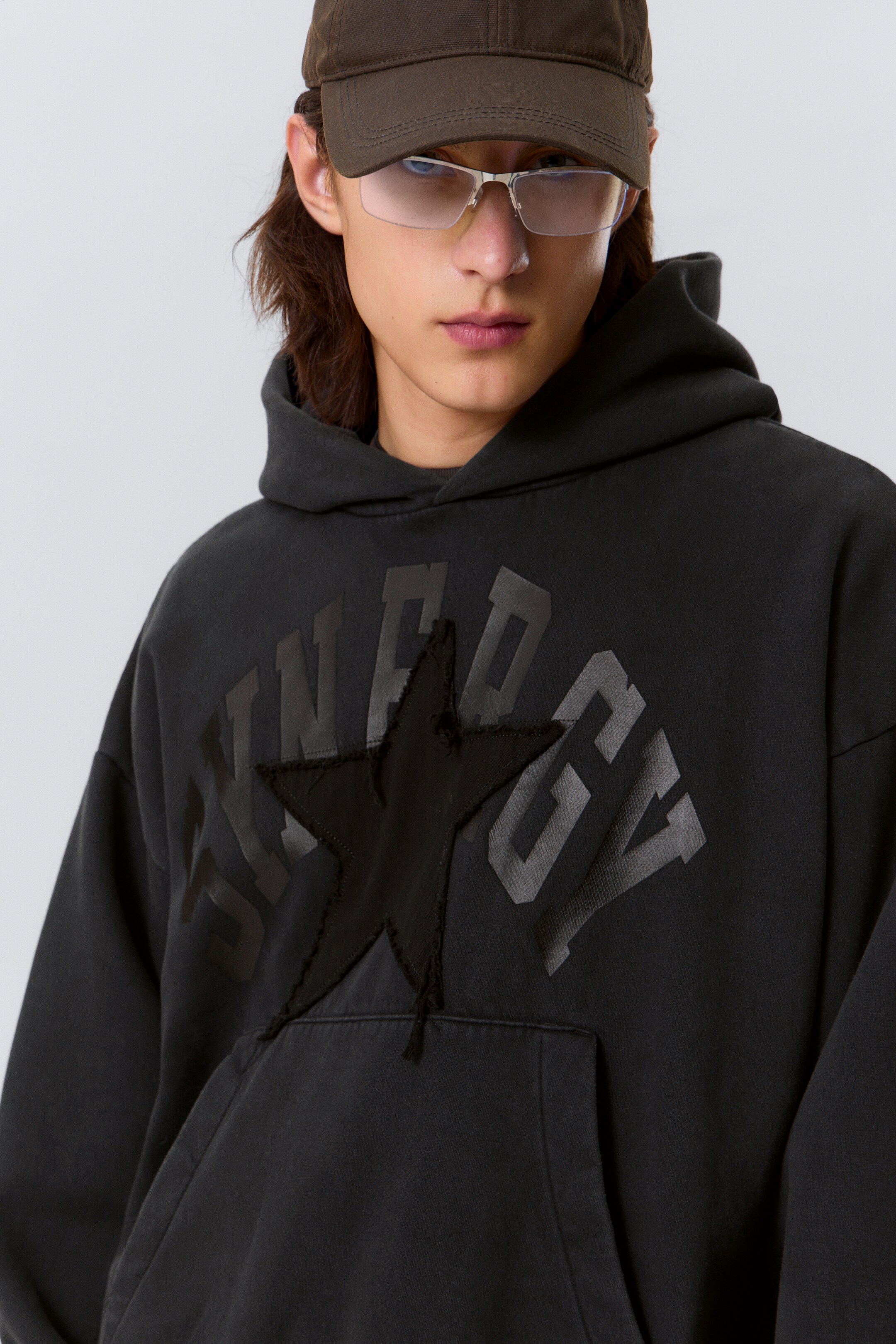 View larger image: A dark grey, washed-effect hoodie is being presented from a slight front-quarter angle. It is featuring large, dark grey graphic lettering across the chest, partially obscured by a prominent black star appliqué with distressed, frayed edges. A spacious kangaroo pocket is visible at the lower front.