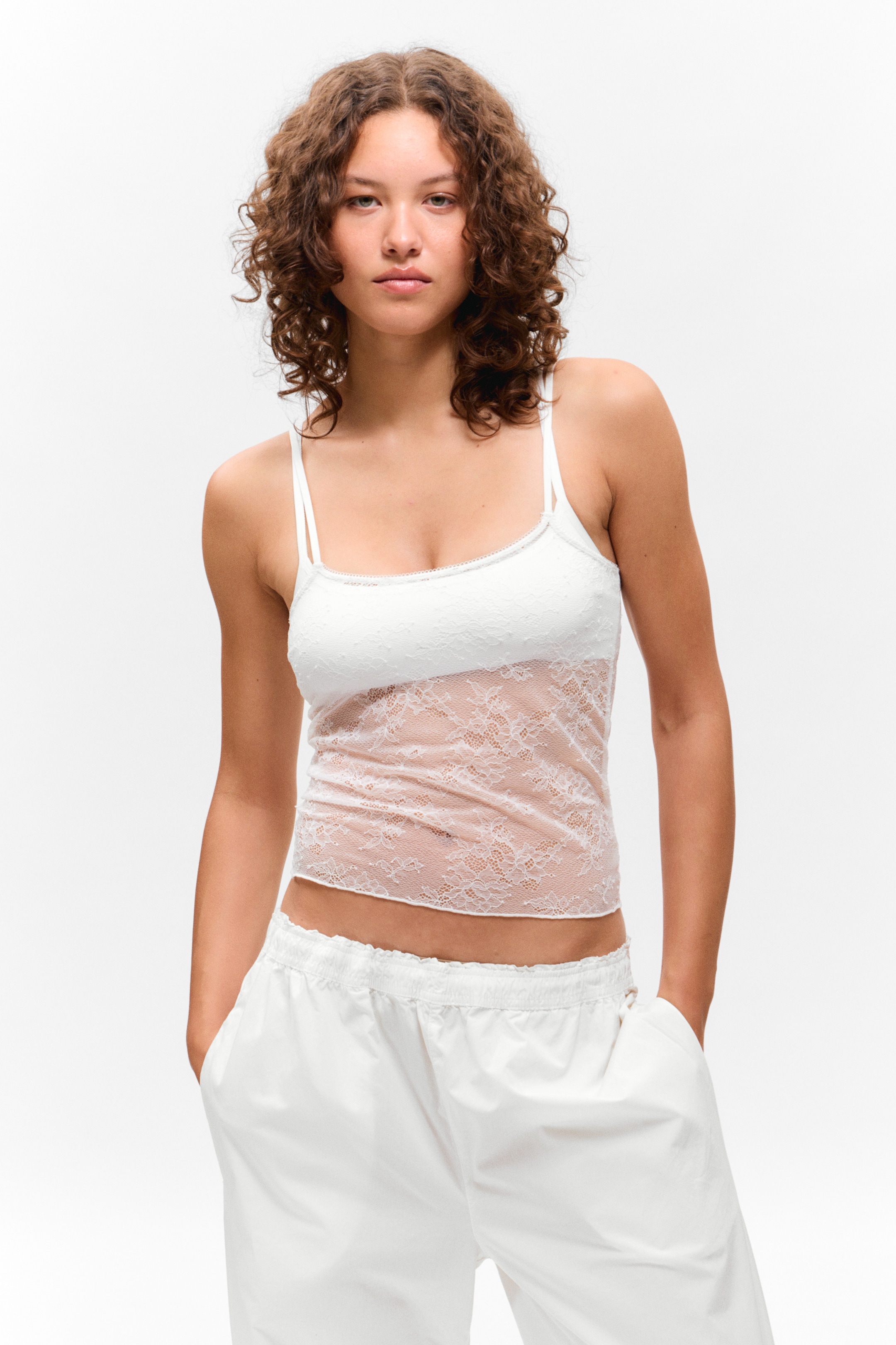 Ver imagen más grande: Se presenta una camiseta de encaje blanco, con un cuerpo de encaje floral transparente, dobles tirantes finos y un delicado ribete festoneado a lo largo del escote y el dobladillo. Está superpuesta sobre una camiseta blanca sólida.