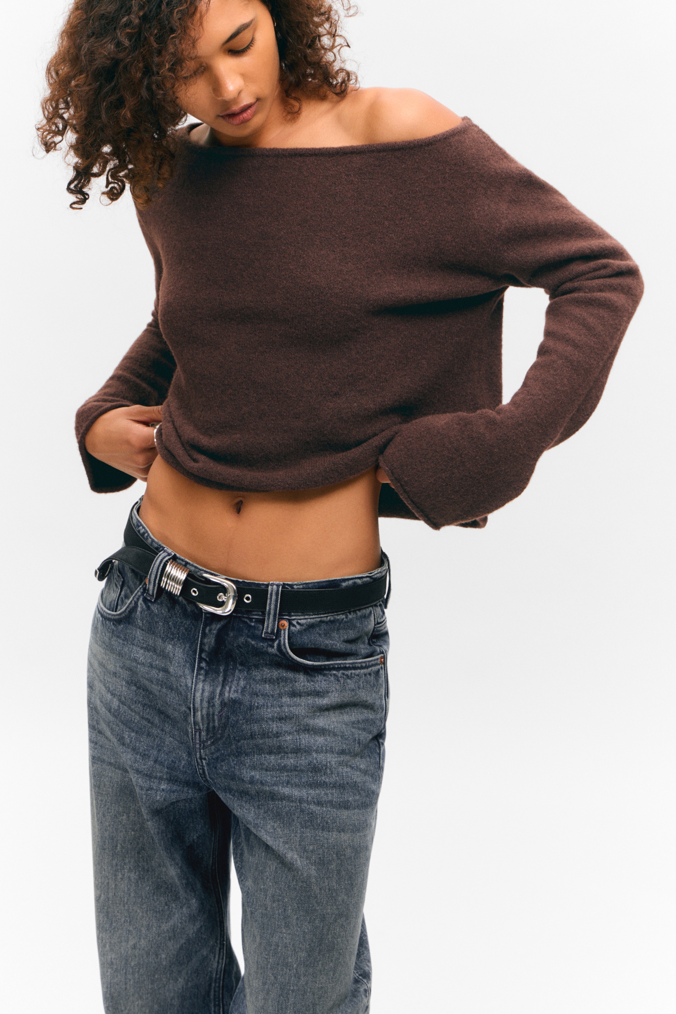 View larger image: A brown, off-the-shoulder sweater with a boat neckline and wide-cuff sleeves is being presented front-facing, appearing slightly cropped. It is styled over dark wash, relaxed-fit jeans, which are secured with a black belt featuring a silver-toned, ornate buckle.