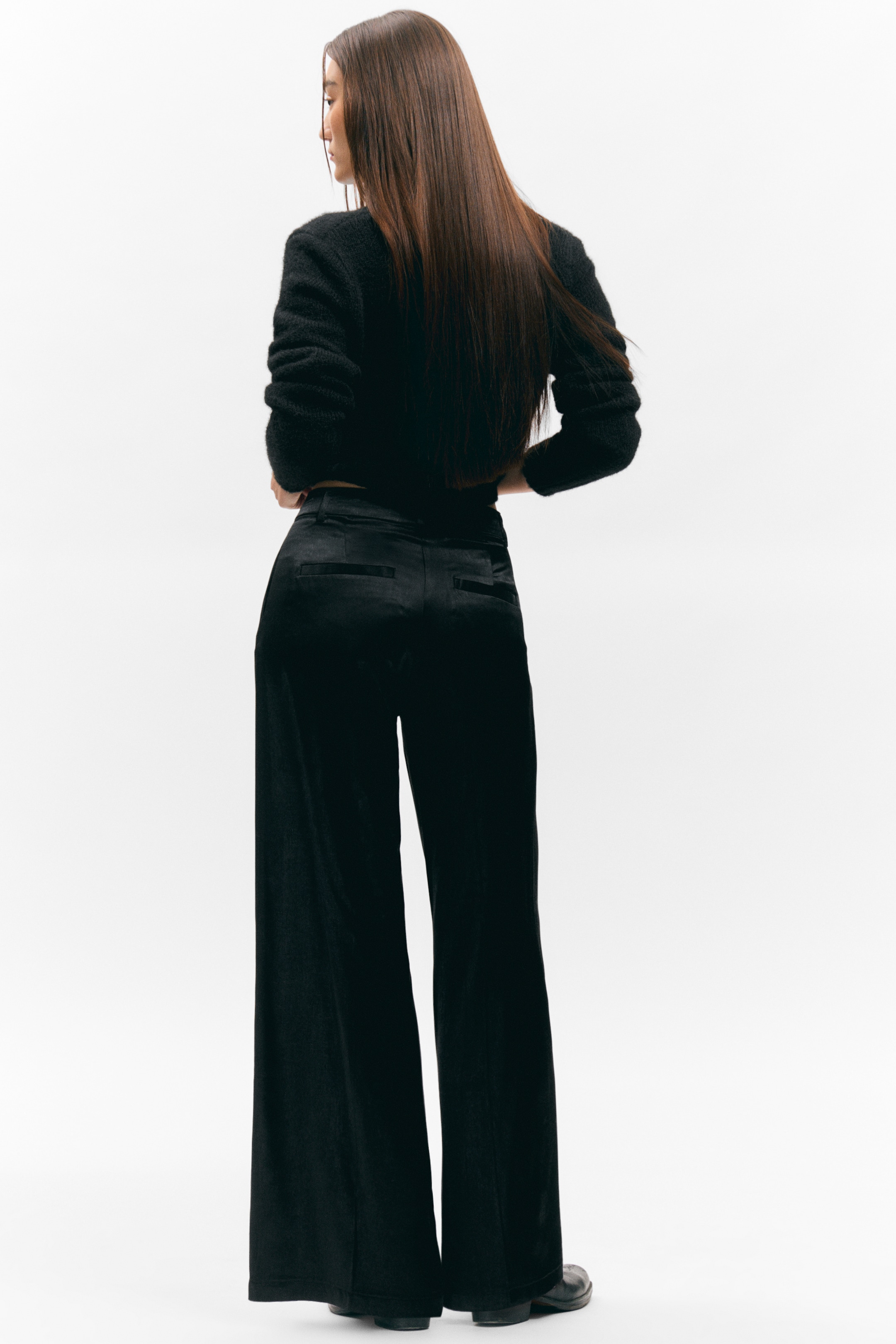 Agrandir l'image: Un pull noir raccourci avec un tricot texturé et des manches longues, présenté de dos et incliné légèrement vers la gauche. Il est porté avec un pantalon noir à jambes larges, qui présente un tissu lisse, semblable à du satin et des poches passepoilées arrière visibles.