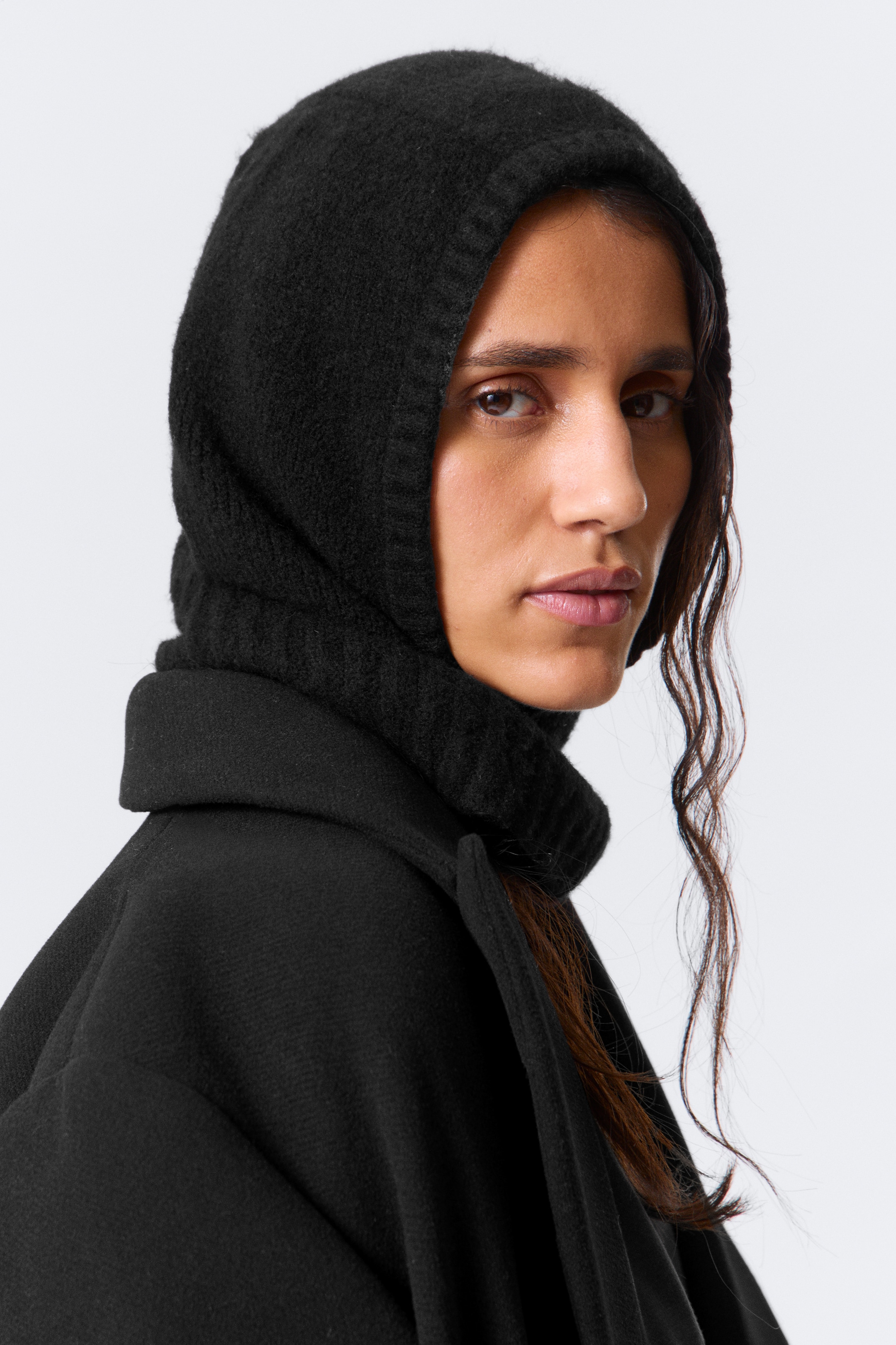 Grösseres Bild ansehen: Eine schwarze, gestrickte Balaclava-ähnliche Kapuze bedeckt den Kopf und den Hals und weist eine flauschige Textur am Kopf und eine gerippte Textur um die Gesichtsöffnung auf. Diese Kapuze ist über einem schwarzen Mantel geschichtet, der ein glattes, gefilztes Gewebe und einen Stehkragen aufweist und aus einem leichten Seitenwinkel gezeigt wird.
