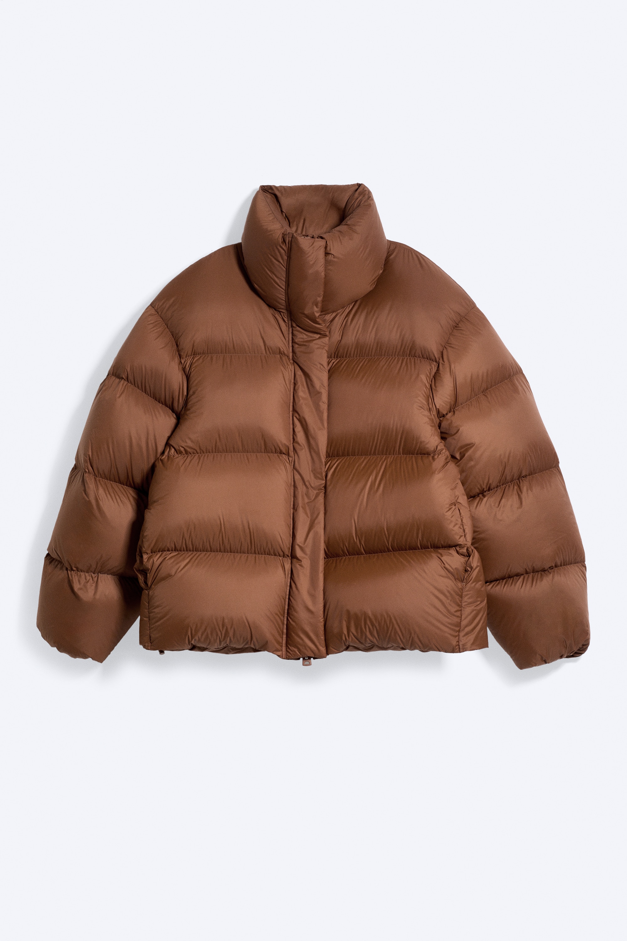 O jachetÄ puffer maro, supradimensionatÄ, cu guler Ã®nalt Èi matlasare orizontalÄ voluminoasÄ, este purtatÄ de modelul aflat Ã®n profil. Jacheta este asortatÄ cu blugi denim cu croialÄ largÄ, efect uzat, Èi o ÈapcÄ cu bordurÄ din blanÄ artificialÄ.