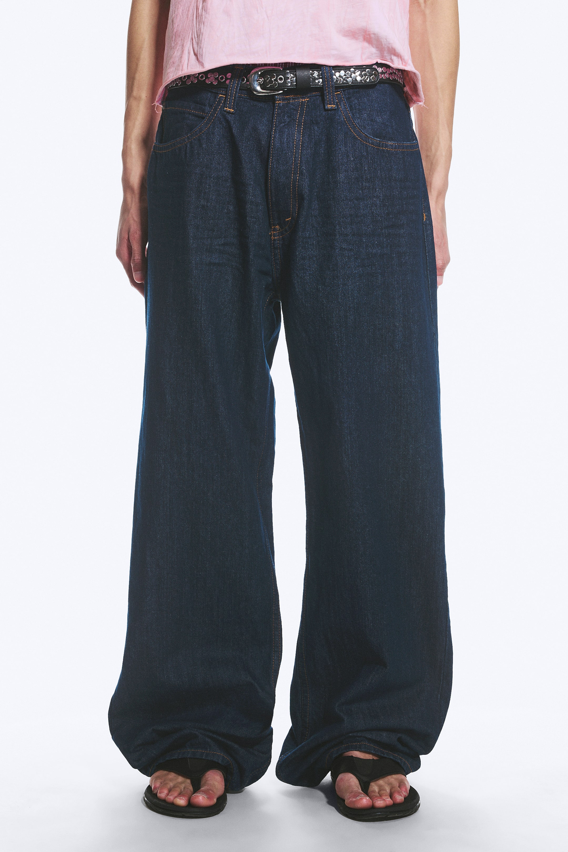 Agrandir l'image: Jean en denim délavé foncé avec une coupe ample et jambes larges, présenté de face. Le jean est maintenu à la taille par une ceinture noire cloutée, et des coutures contrastées sont visibles autour des poches avant et de la braguette.