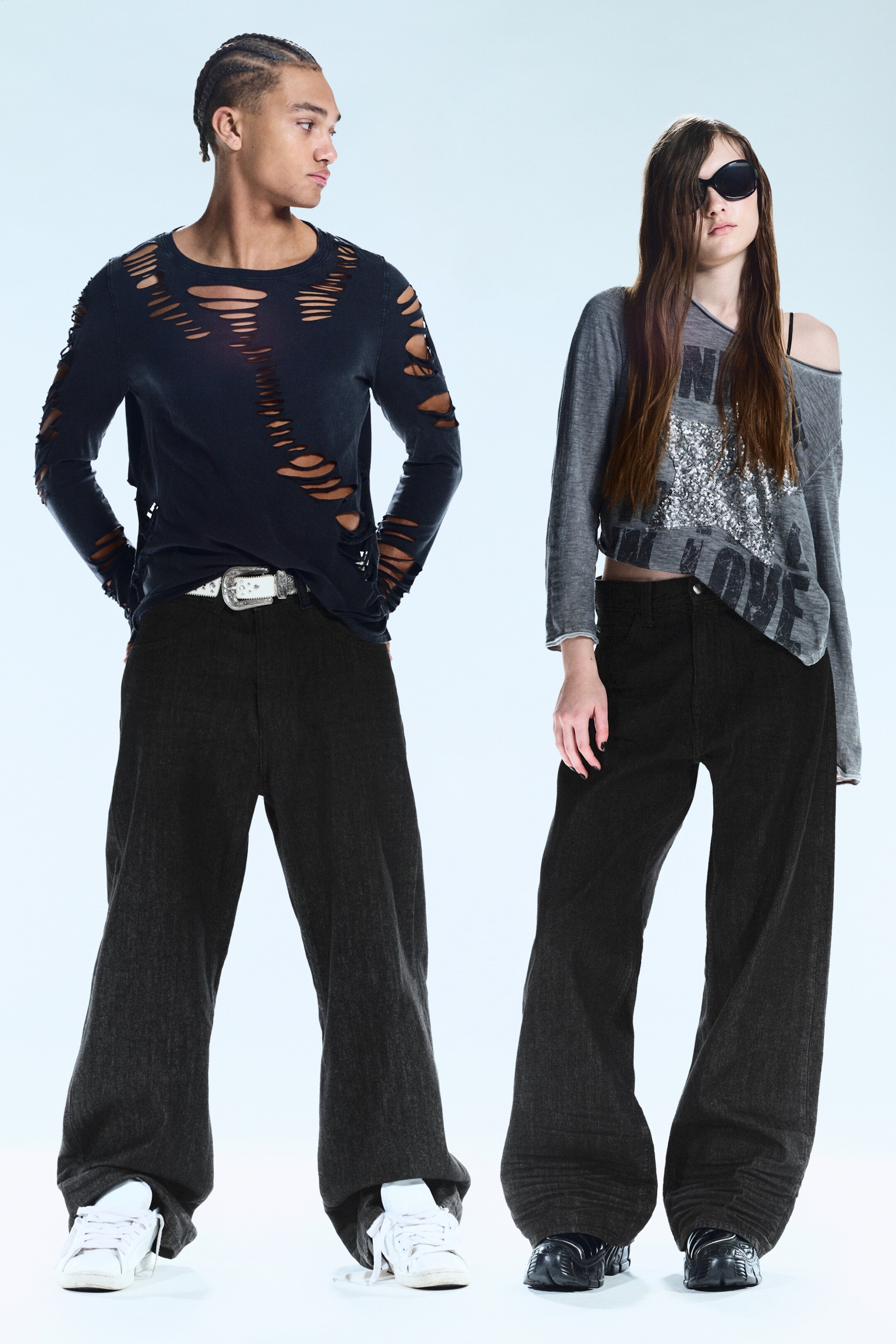 Zobacz większy obraz: Czarna koszula z długim rękawem z szerokimi poziomymi przetarciami jest prezentowana w zestawieniu z ciemnoszarymi spodniami z szerokimi nogawkami oraz białym paskiem z ozdobną srebrną klamrą. Również prezentowana jest szara bluzka z długim rękawem, odsłaniająca ramiona, z cekinowym wzorem graficznym, zestawiona z ciemnoszarymi spodniami z szerokimi nogawkami.