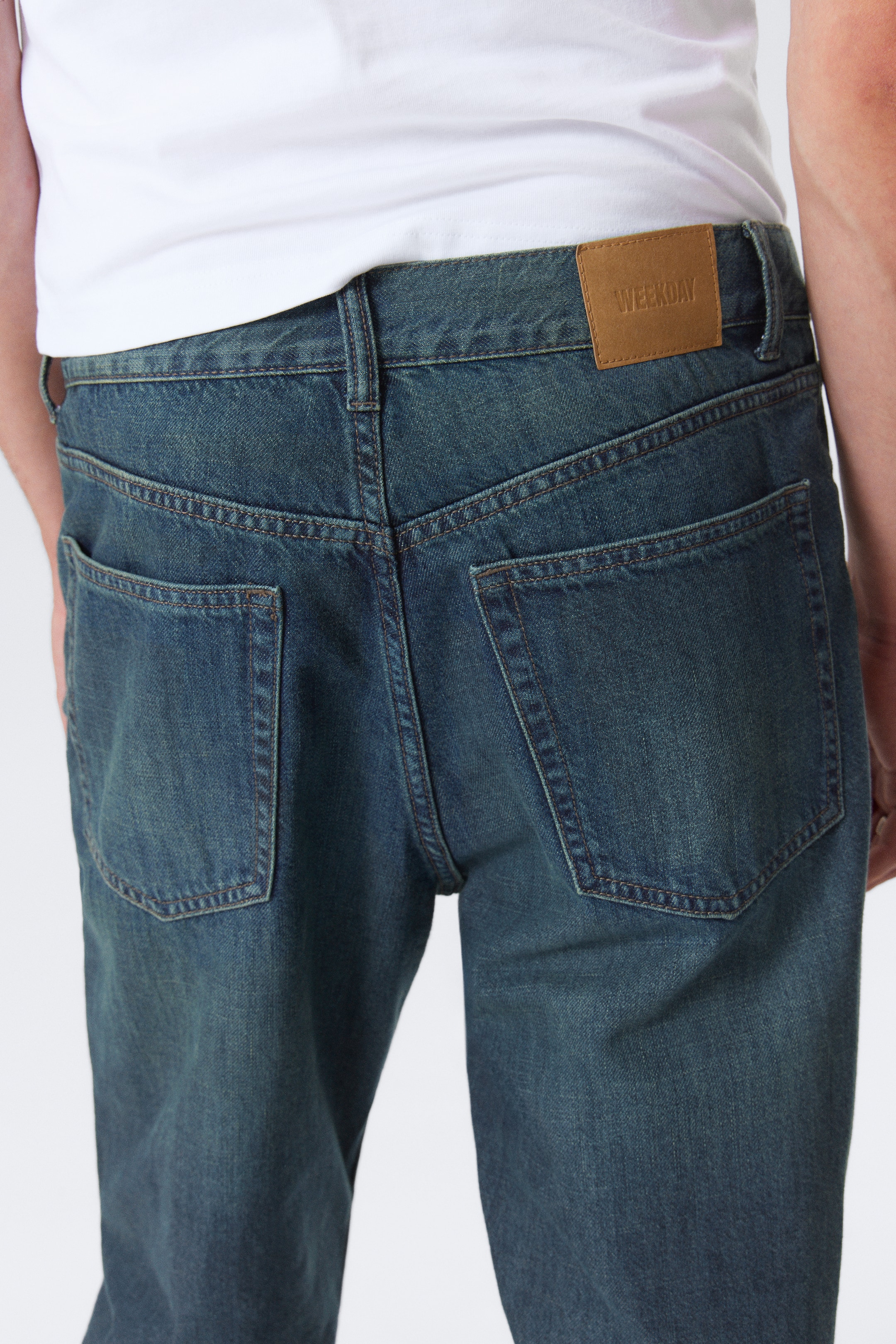 Visa större bild: Mörkblå denimjeans presenteras från en bakre vinkel, som visar två stora påsydda fickor med framträdande orange kontrastsömmar. En brun märkeslapp, präglad med 'WEEKDAY', är fäst på höger sida av midjebandet, och en vit t-shirt bärs tillsammans med jeansen.