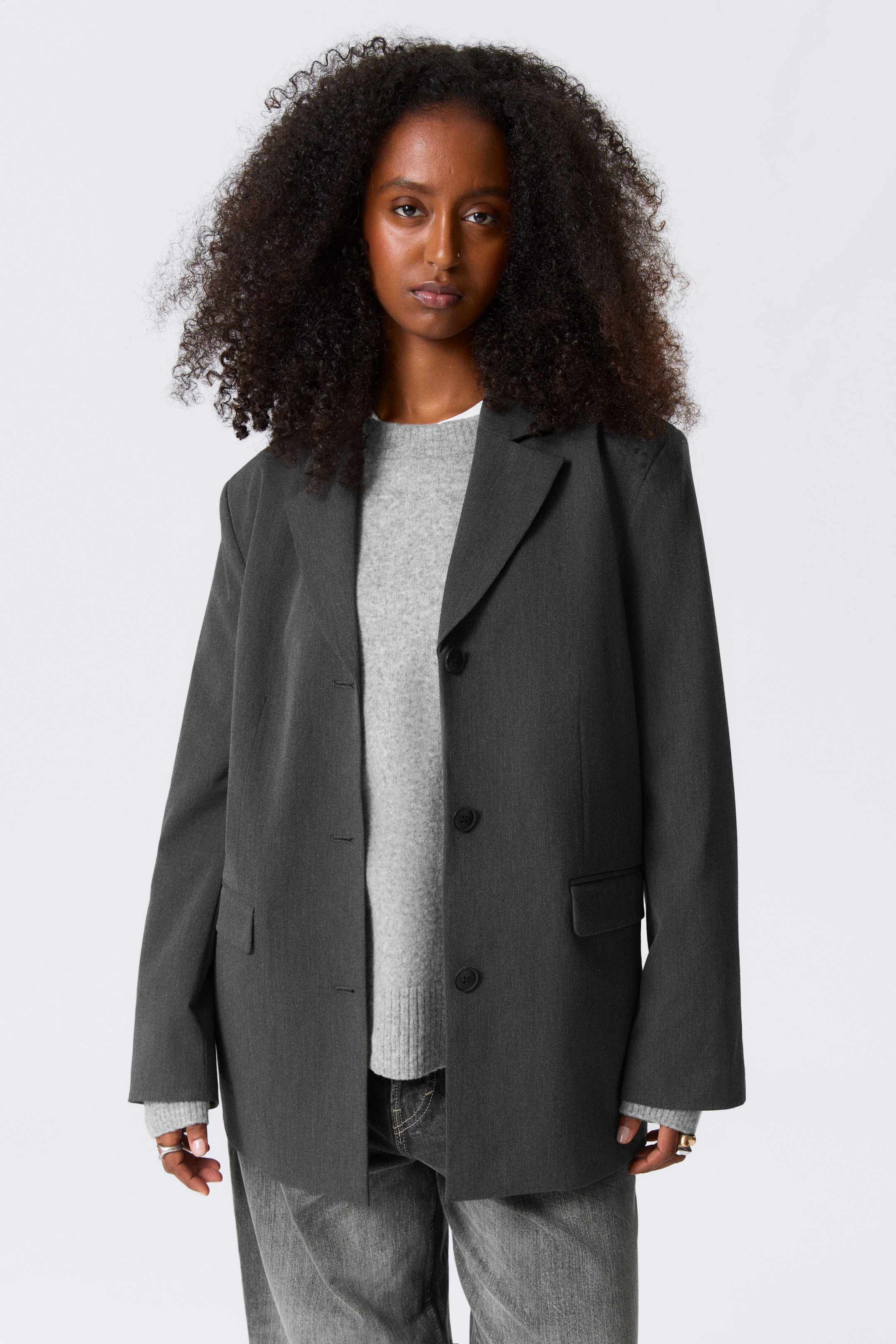 Visualizza immagine più grande: Un blazer monopetto grigio scuro viene indossato, rivolto in avanti, presentando una vestibilità oversize. Presenta revers a dente, una chiusura a tre bottoni e due tasche con patta, ed è stratificato su un maglione girocollo grigio chiaro.