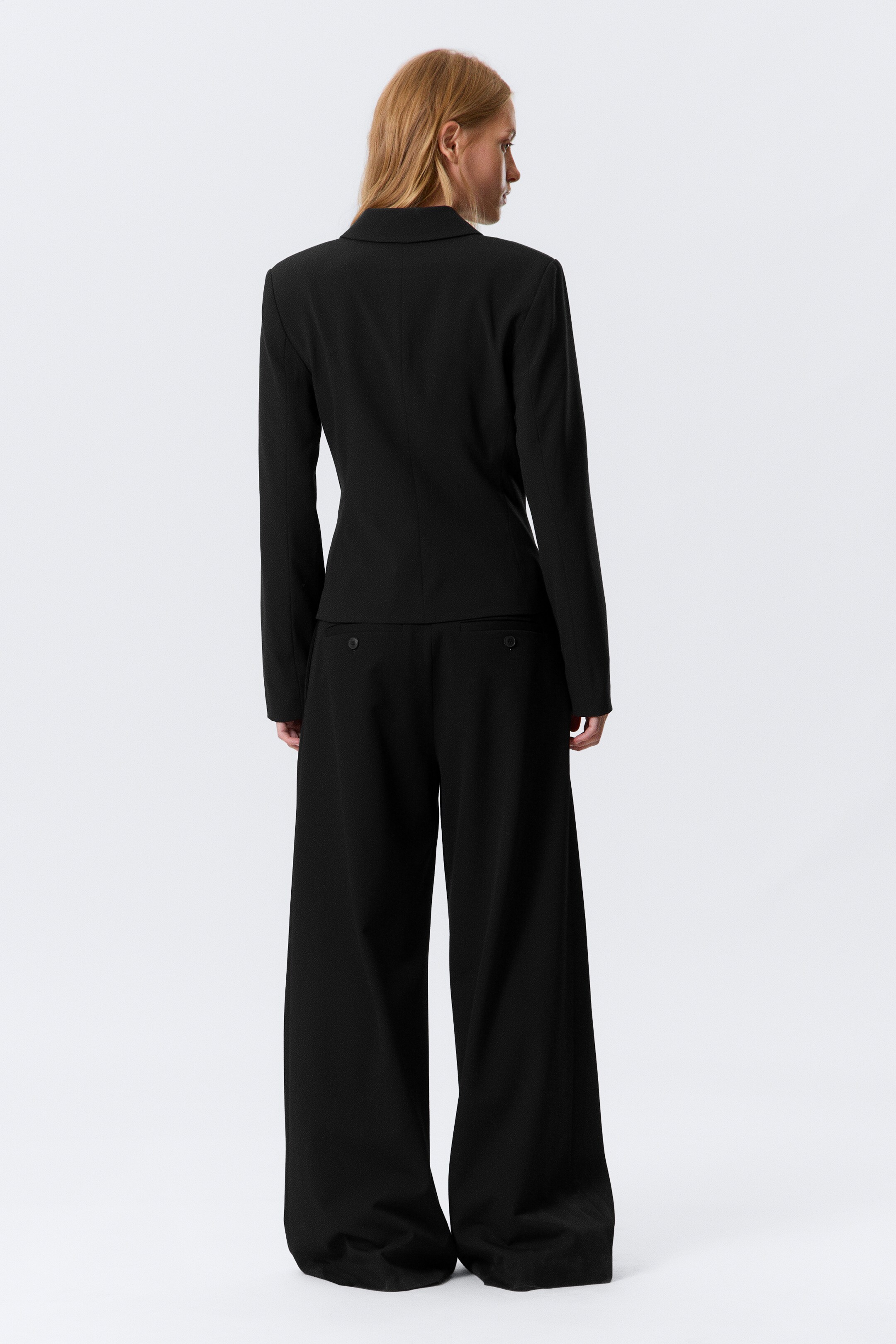 Agrandir l'image: Un tailleur-pantalon noir est présenté de dos, avec un blazer ajusté et un pantalon à jambes larges. Le blazer présente des manches longues et une coupe ajustée et courte à la taille, tandis que le pantalon est conçu avec une silhouette fluide et deux poches passepoilées boutonnées à l'arrière.