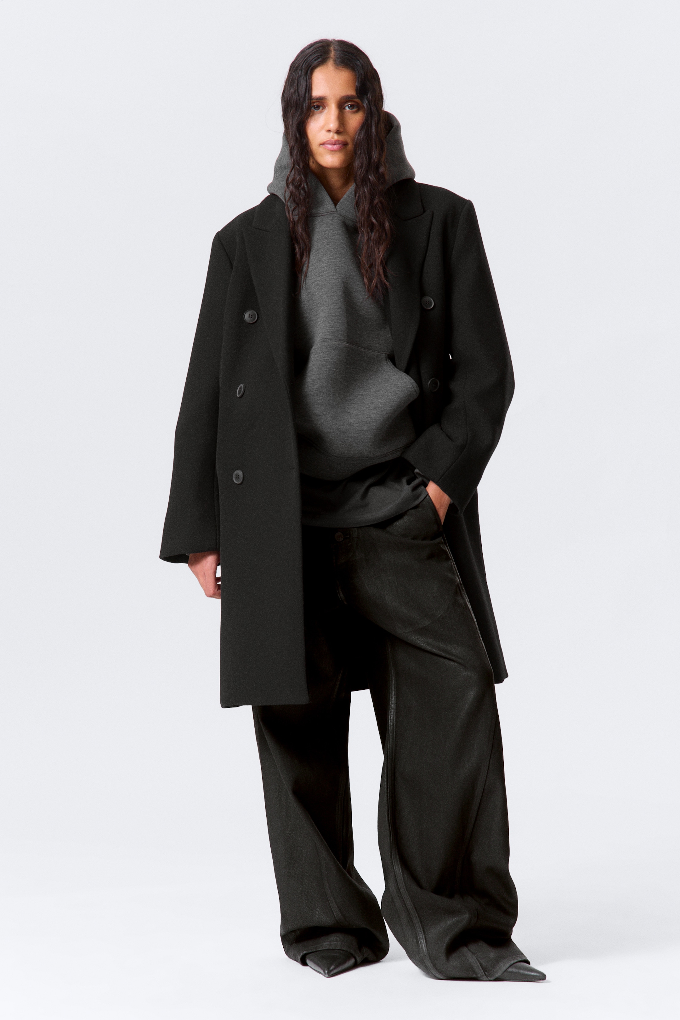 Ver imagem maior: Um casaco preto de trespasse com lapelas largas e quatro botões visíveis está a ser mostrado de frente, aberto sobre um hoodie cinzento escuro com bolso canguru. O casaco oversized cai até ao meio da coxa, e calças largas e escuras com costuras verticais acumulam-se sobre sapatos pretos de bico fino.