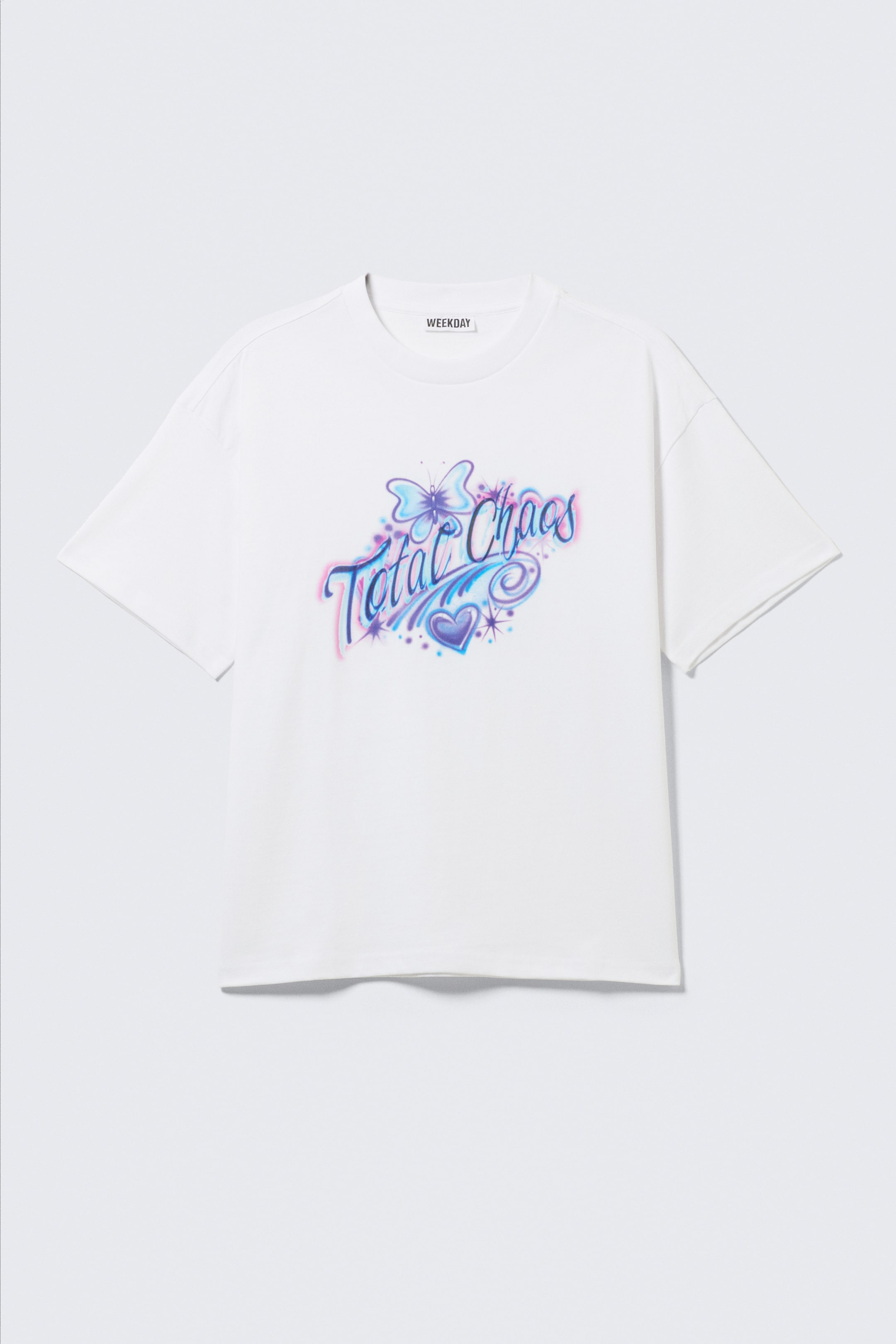 Zobraziť väčší obrázok: Biele, voľne padnúce tričko je zobrazené spredu, s výraznou grafickou potlačou "Total Chaos" s motívom motýľa a srdca v odtieňoch modrej, fialovej a ružovej. Navrhnuté je s okrúhlym výstrihom, obalenými švami, spustenými ramenami a dvojitým prešitím na lemoch.