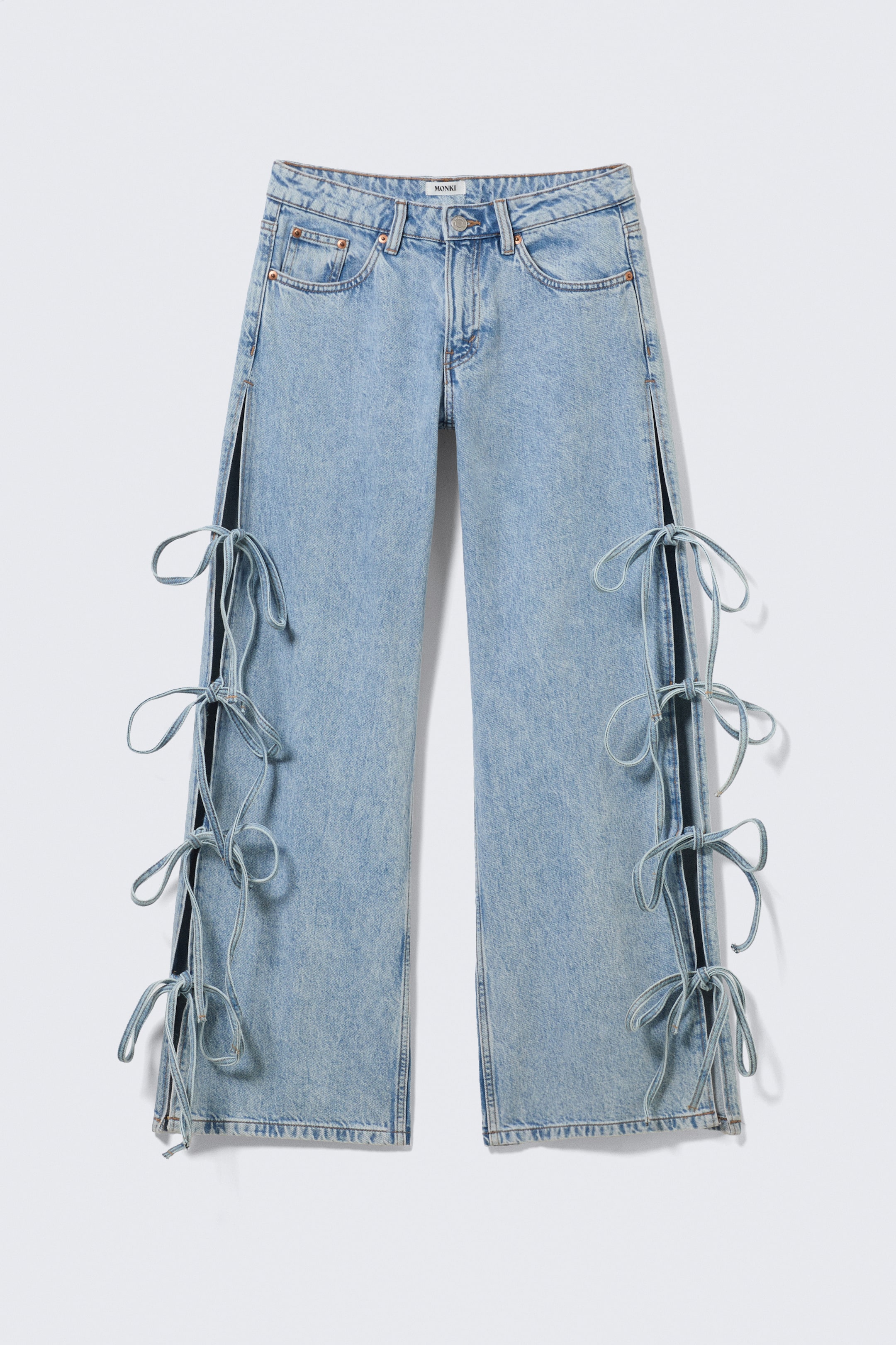 Agrandir l'image: Jean en denim délavé bleu clair, arborant une étiquette de marque "MONKI" sur la ceinture, présenté de face avec une coupe droite. Des fentes latérales ouvertes descendent le long des coutures extérieures, révélant un tissu plus sombre en dessous, et sont maintenues par plusieurs liens en denim façonnés en nœuds.