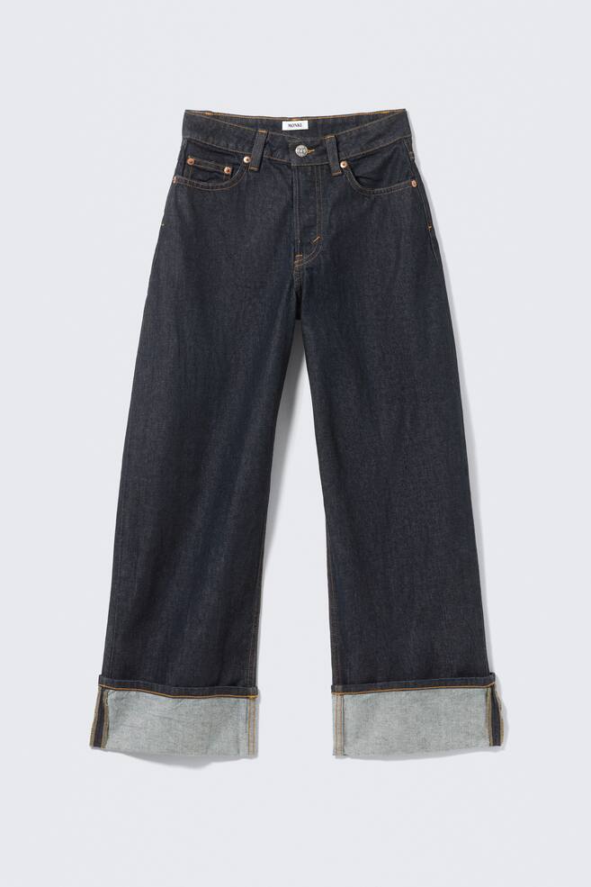 Donker indigo jeans liggen VOORAAN, met een lage taille, losse pasvorm en super wijde pijpen. Ze hebben een klassiek vijfzakkenontwerp met koperen klinknagels, contrasterende bruine stiksels en een ritssluiting, terwijl de onderzomen zijn opgerold, waardoor het lichtere denim interieur zichtbaar wordt. - 1