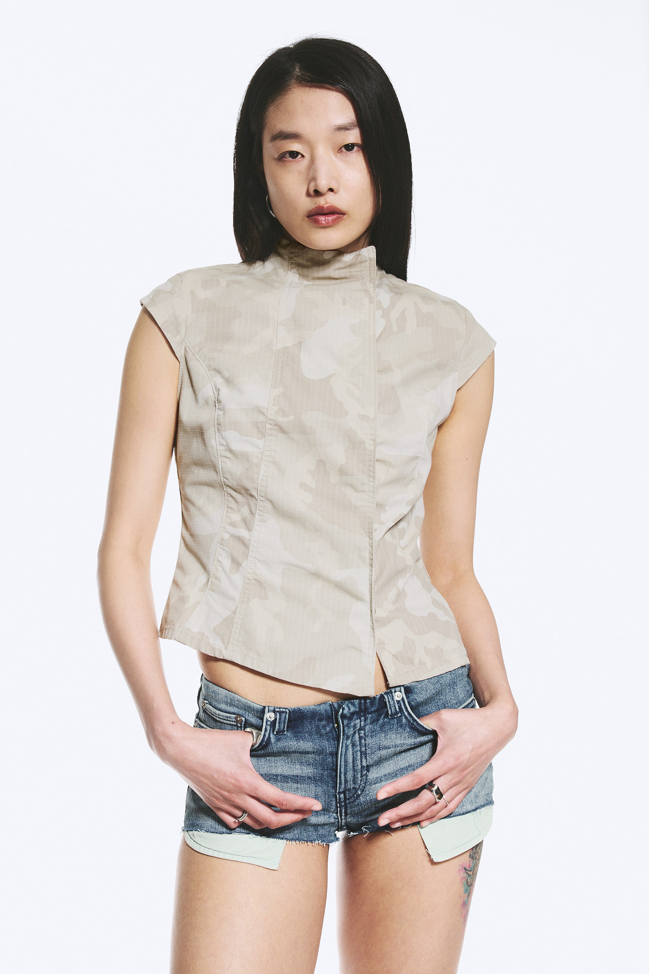 Ver imagen más grande: La modelo lleva un top ajustado con un ligero estampado de camuflaje. Presenta un cuello simulado, mangas cortas, detalles de costura curvada para una silueta estructurada, un cierre frontal oculto y un dobladillo asimétrico.