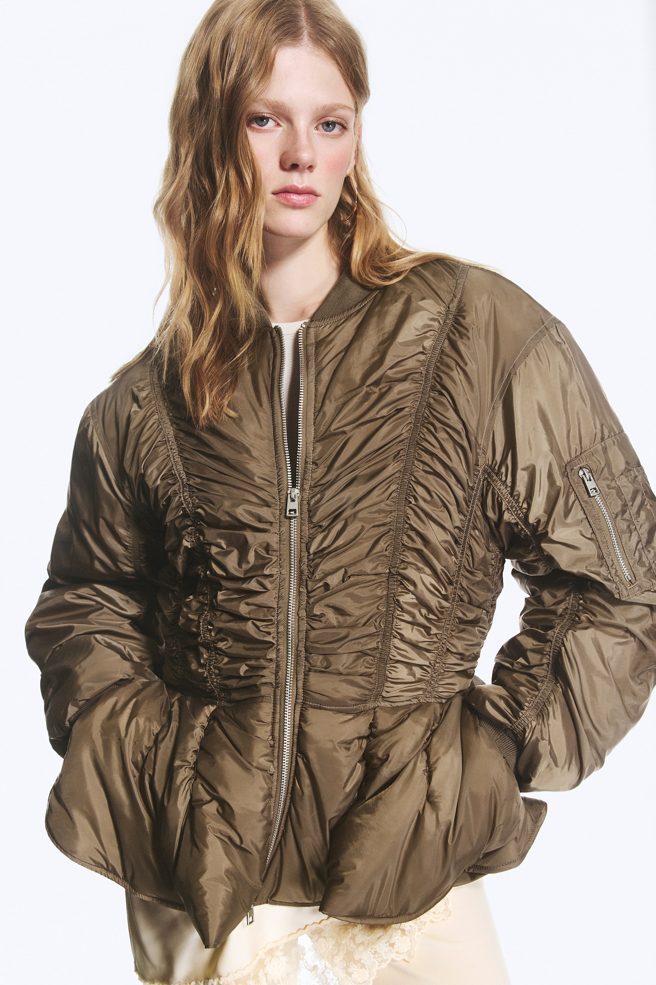 Agrandir l'image: Un blouson bomber ajusté de couleur olive-brun est porté par le mannequin, avec des coutures froncées sur toute la surface, une fermeture éclair frontale à double sens et une poche zippée sur la manche. Le blouson présente un ourlet à basque, et le mannequin glisse ses mains dans les poches latérales en couture.