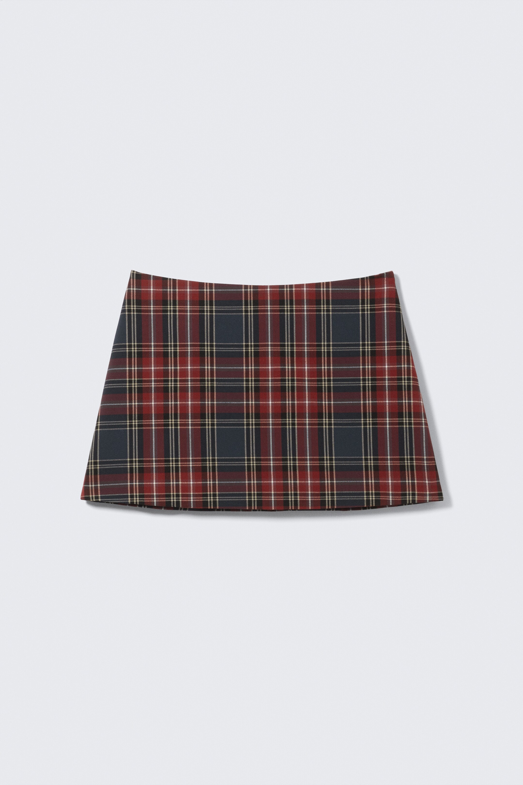 View larger image: A mini skirt is lying flat, showcasing its front. It is featuring a plaid pattern in dark blue, red, black, white, and yellow, designed with a straight waist and a slight A-line shape that ends in a straight hem.