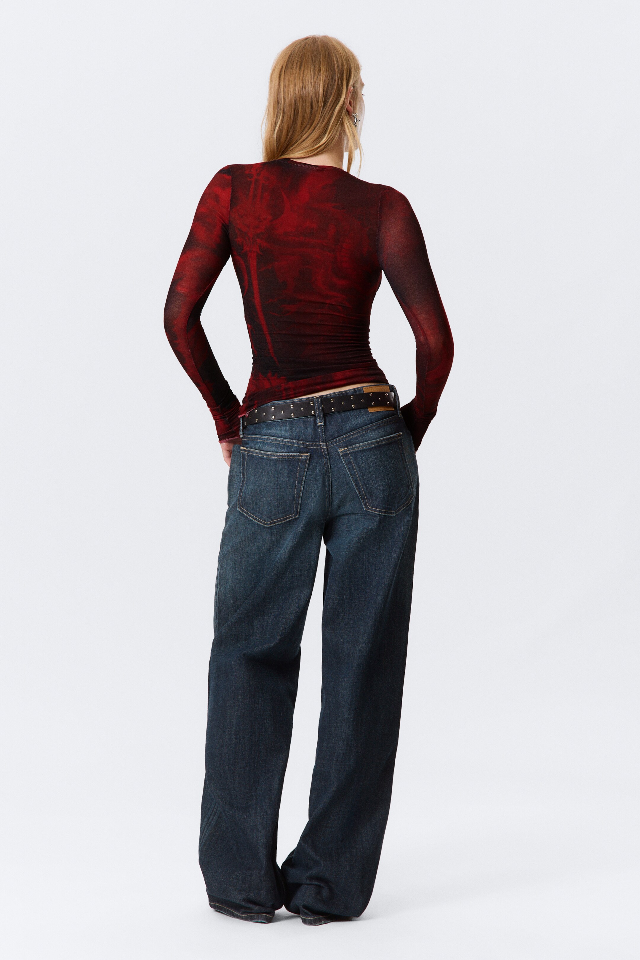 Grotere afbeelding bekijken: Een slim-fit top met lange mouwen, met een rood en zwart bedrukt ontwerp, wordt gedragen door het model. Het topje is gestyled met donker gewassen wijde jeans en een zwart riem met studs.