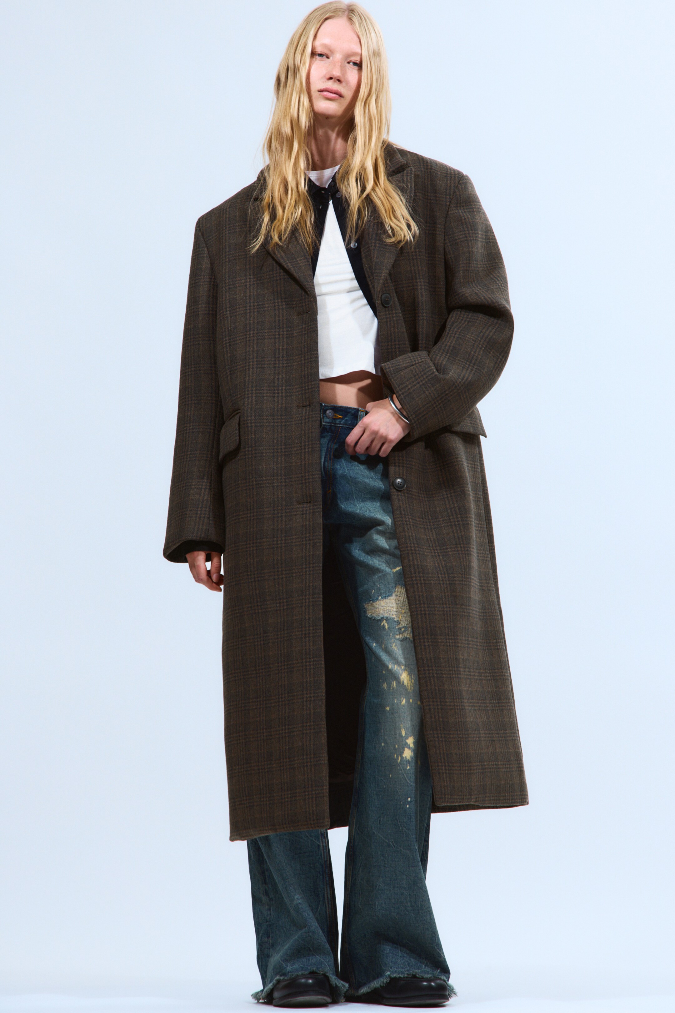 View larger image: A long, dark brown coat with a subtle plaid pattern is being presented from a slightly angled front view, showcasing its oversized fit. It is featuring notch lapels, a single-breasted button closure, and two visible flap pockets, with its hemline extending to just above the ankles.