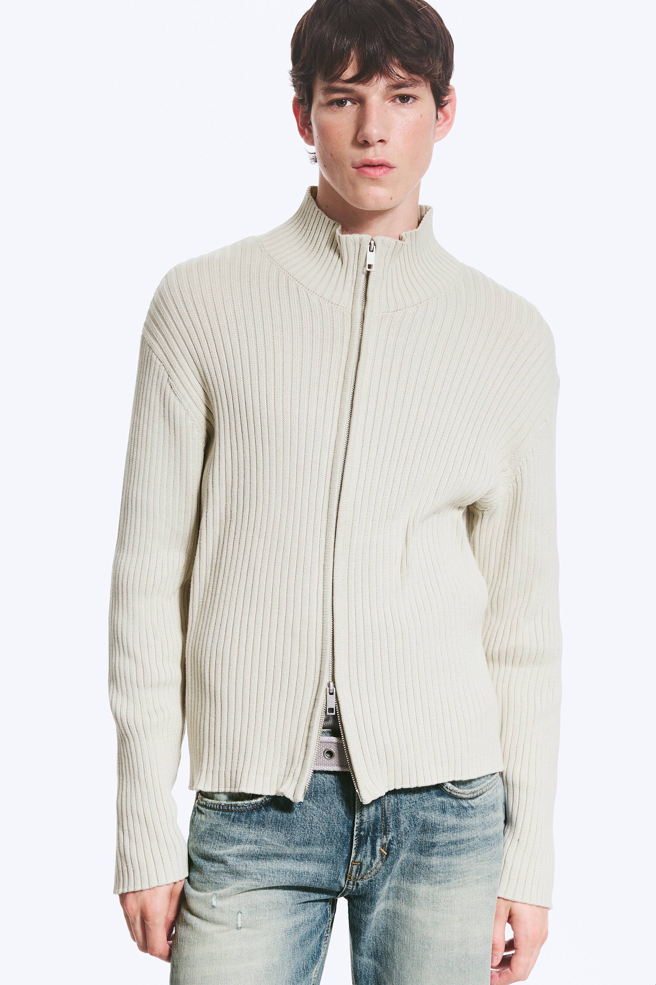 Visualizza immagine più grande: Un maglione a coste color écru con zip integrale è indossato, caratterizzato da un collo alto, prominenti coste verticali lungo tutto il corpo e maniche lunghe, e da una zip color argento parzialmente aperta dal basso, che rivela una cintura e uno strato interno.