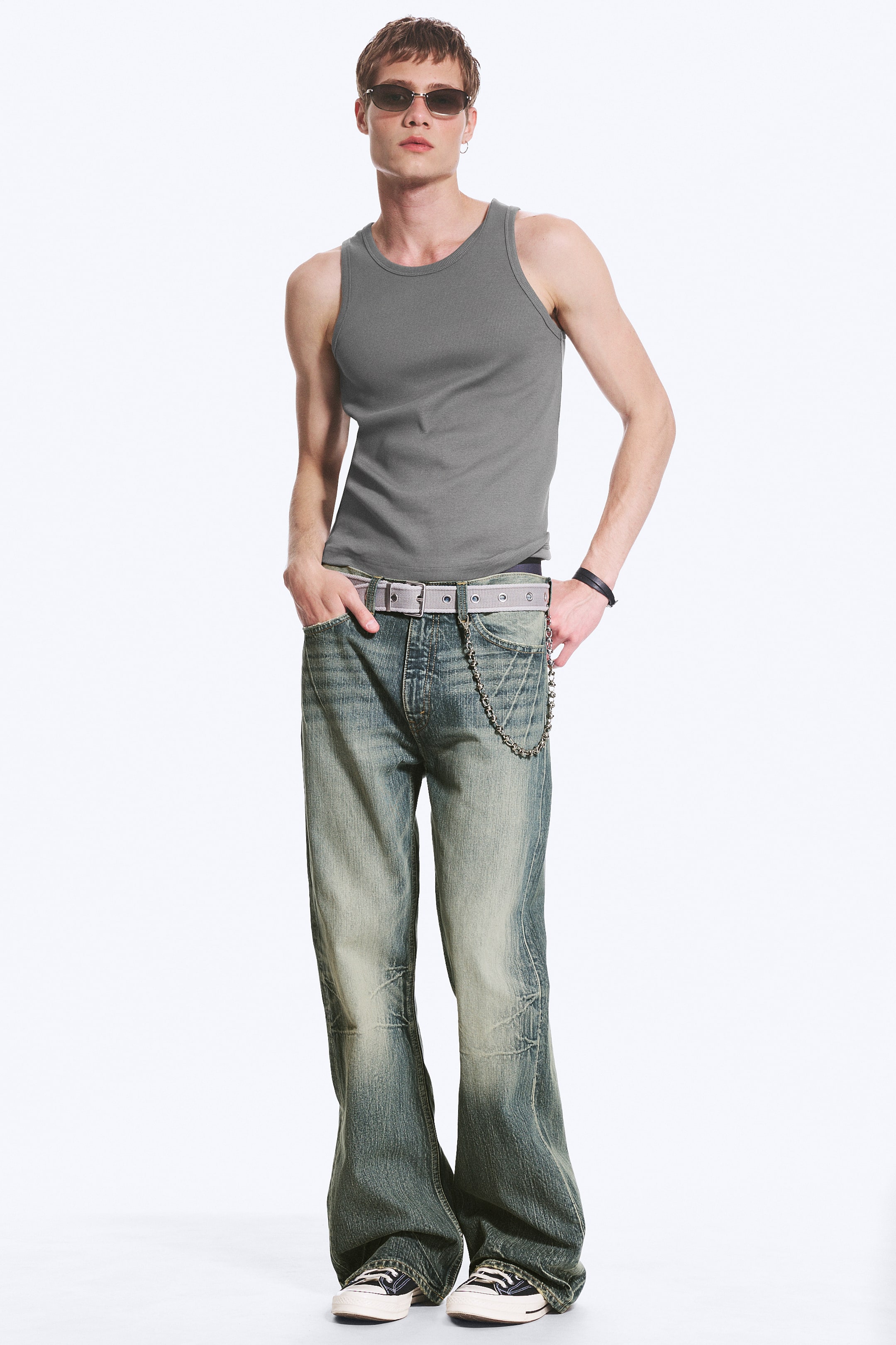 Größeres Bild ansehen: Bootcut-Jeans mit Used-Waschung, die durch eine niedrig sitzende Taille und ein Five-Pocket-Design gekennzeichnet sind, werden vom Model getragen. Ein hellgrauer Gürtel ist durch die Gürtelschlaufen gezogen, und ein Metallketten-Accessoire hängt von einer vorderen Gürtelschlaufe, während die Hände des Models in den vorderen Taschen ruhen.