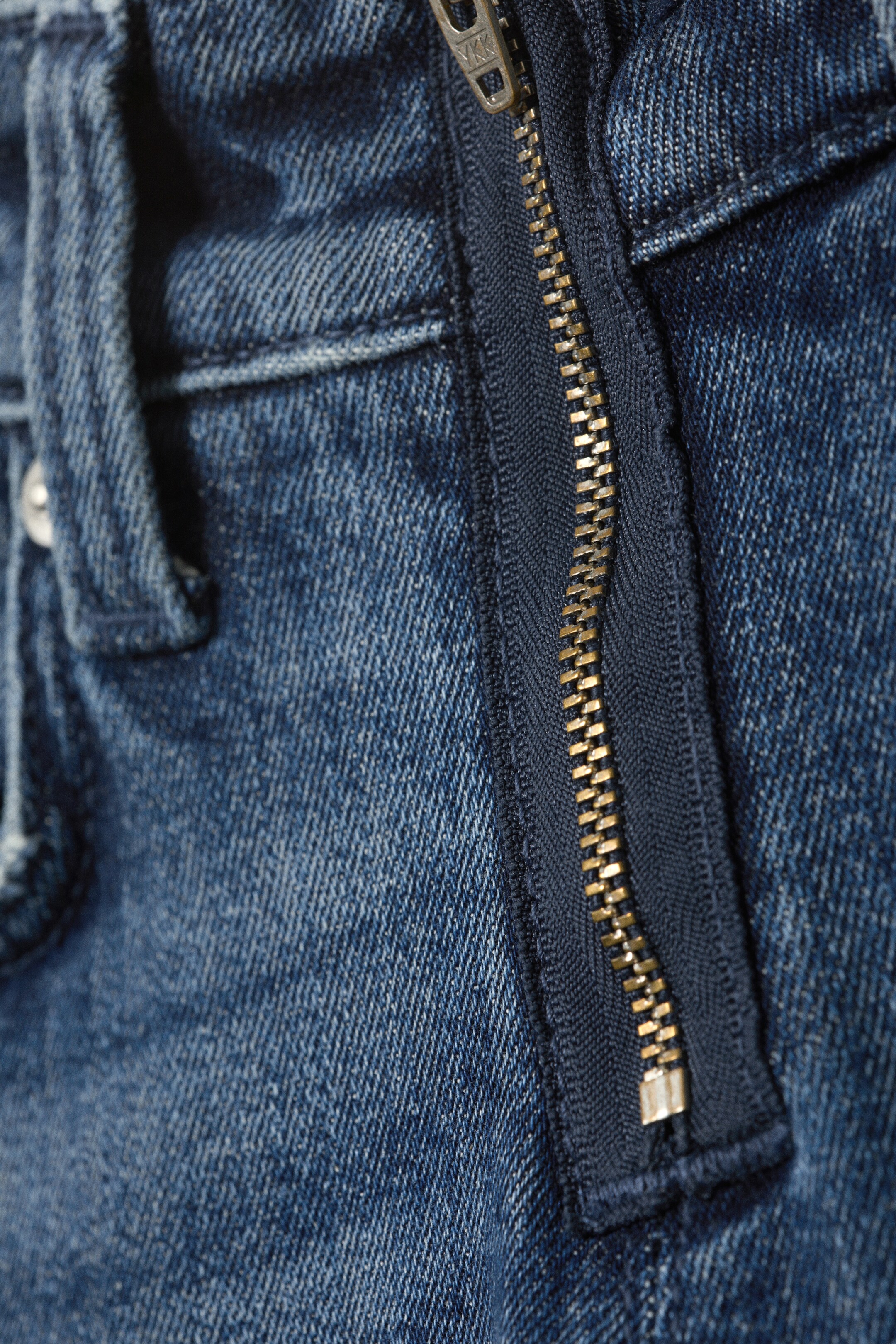 Größeres Bild anzeigen: Eine blaue Denimjeans präsentiert einen teilweise geöffneten metallischen Reißverschluss mit goldfarbenen Zähnen und dunkelblauem Reißverschlussband. Sichtbare Nähte verzieren den Jeansstoff, und eine Gürtelschlaufe erscheint auf der linken Seite.