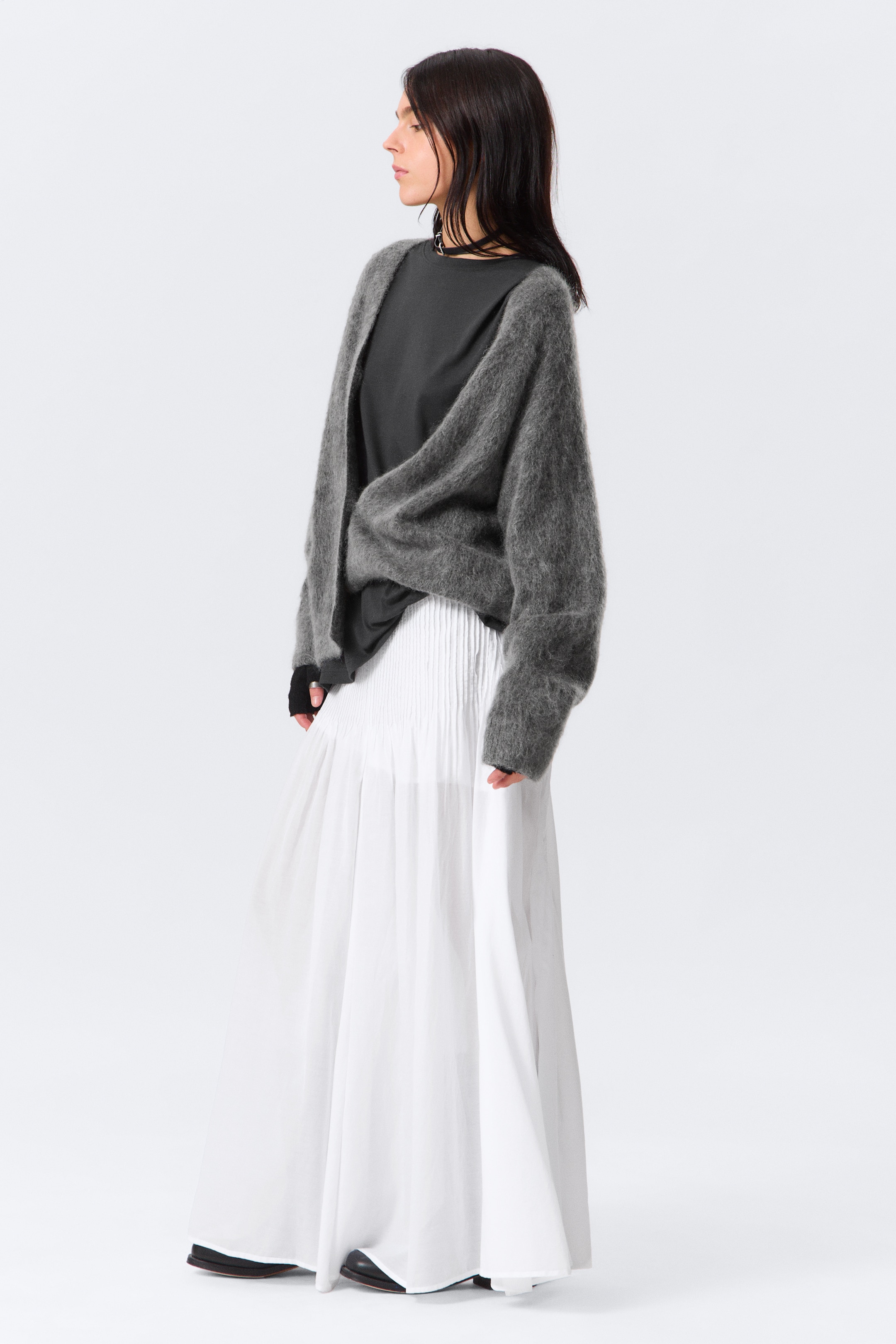 Agrandir l'image: Un cardigan gris foncé et duveteux est porté ouvert sur un haut noir à manches longues, superposé d'une jupe longue plissée blanche. Le cardigan présente une coupe ample et drapée avec des manches longues, tandis que la jupe met en valeur de fins plis à la taille qui se transforment en une longueur complète fluide.