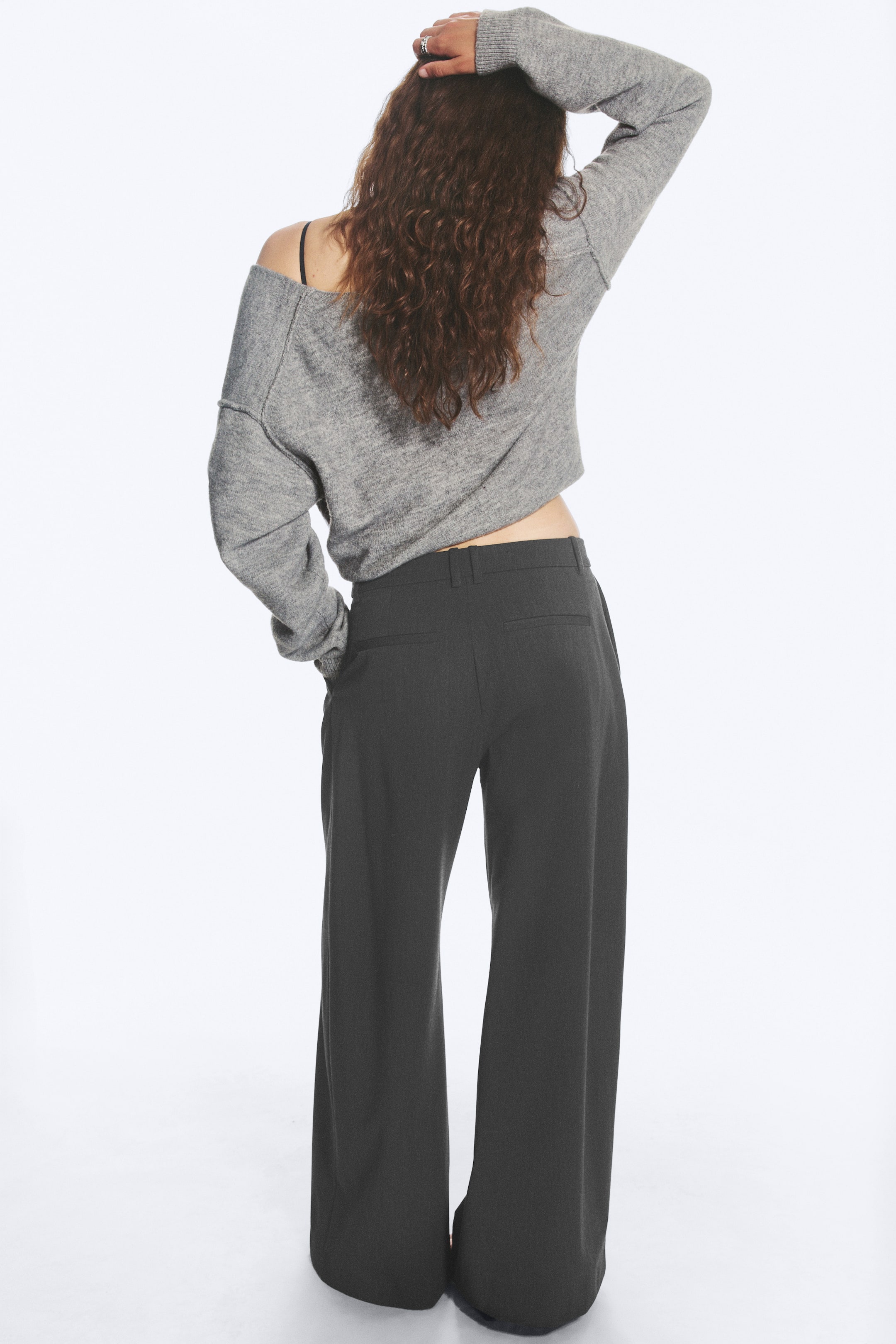 Agrandir l'image: Le mannequin porte un pantalon de costume gris foncé taille basse, doté d’une coupe large, de passants de ceinture et de deux poches passepoilées au dos. Le mannequin est debout, une main glissée dans une poche arrière, avec un pull gris court porté au-dessus, mettant en valeur la taille basse.